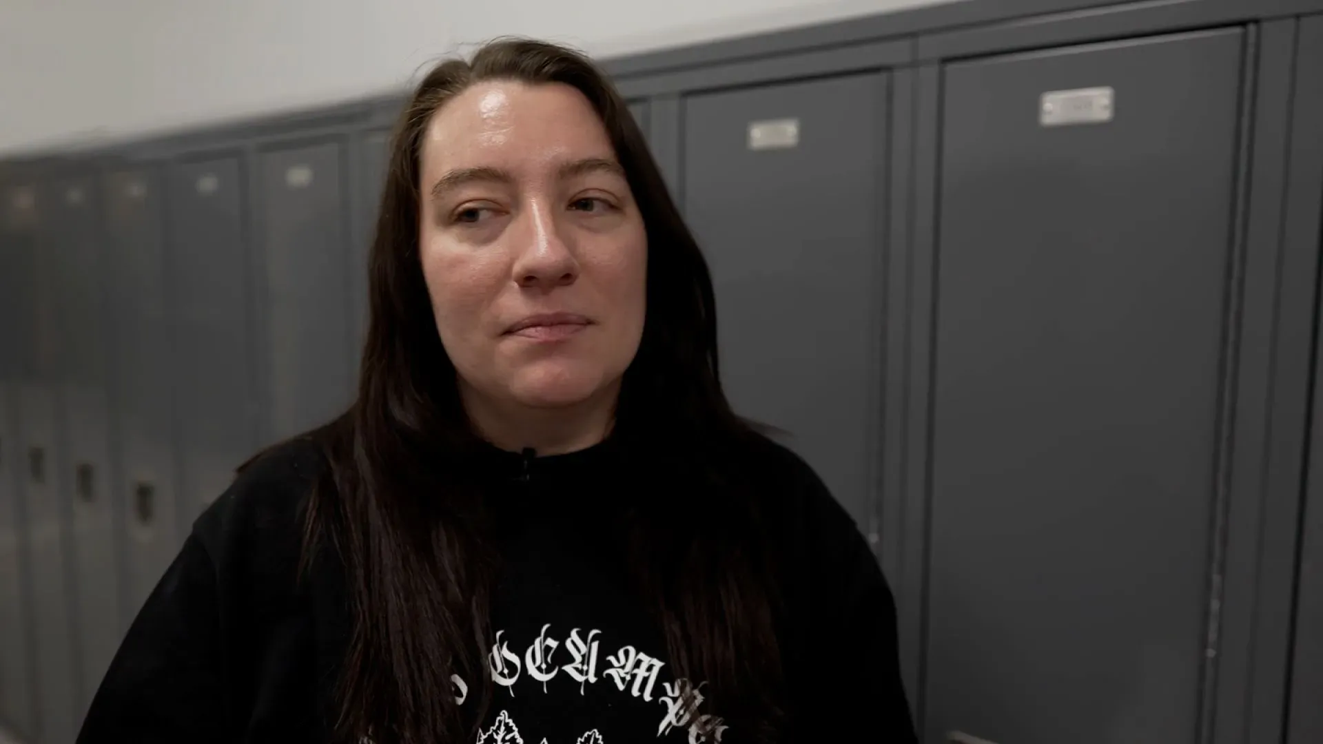 امرأة شابة تتحدث بقلق في ممر مدرسة ثانوية، خلفها خزائن مغلقة، تعكس أجواء الخوف والقلق في مجتمعها بسبب قضايا الهجرة.