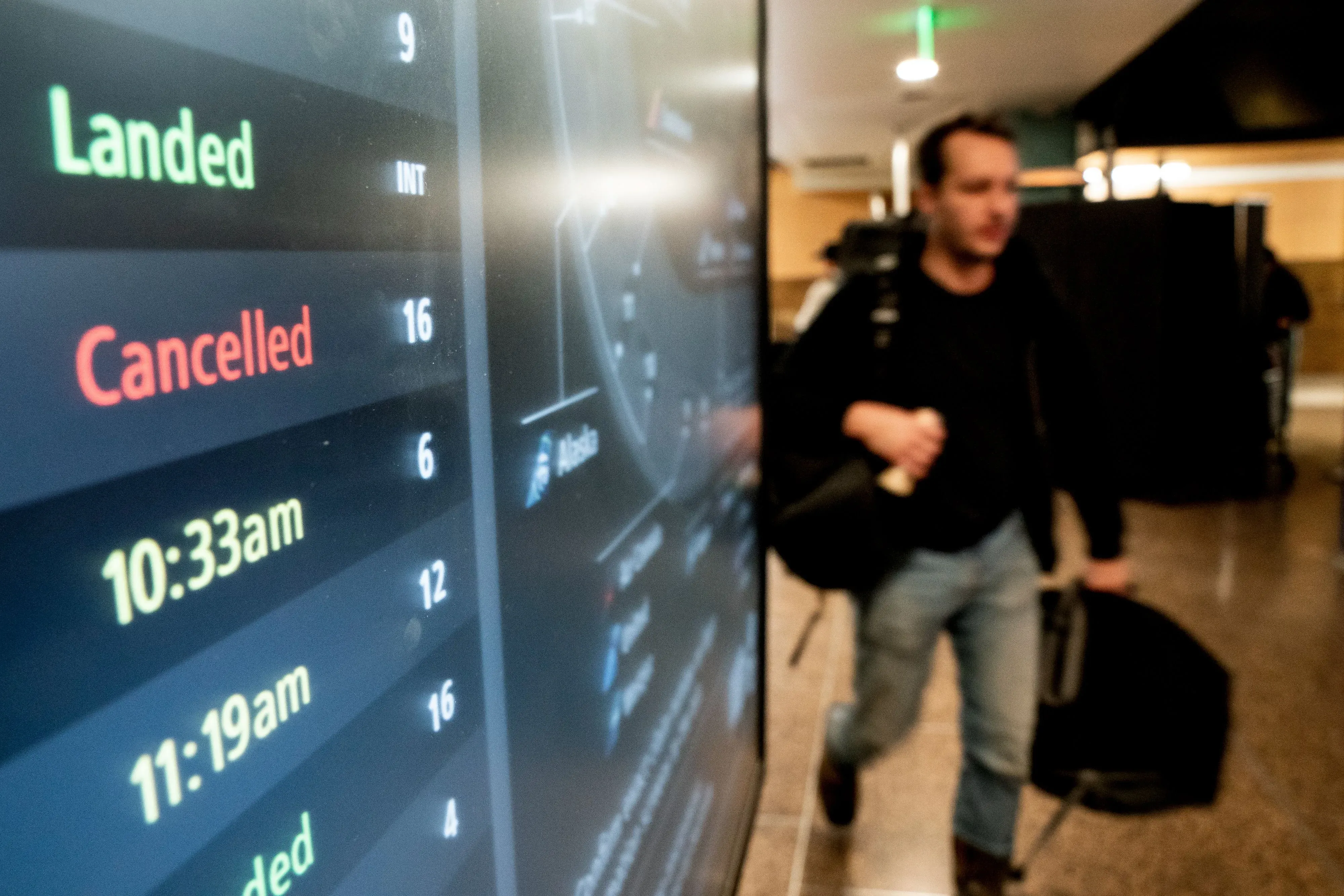 شاشة عرض في مطار تظهر 16 رحلة ملغاة، بينما يسير مسافر يحمل حقيبة، في ظل اضطرابات السفر بسبب نقص موظفي مراقبة الحركة الجوية.