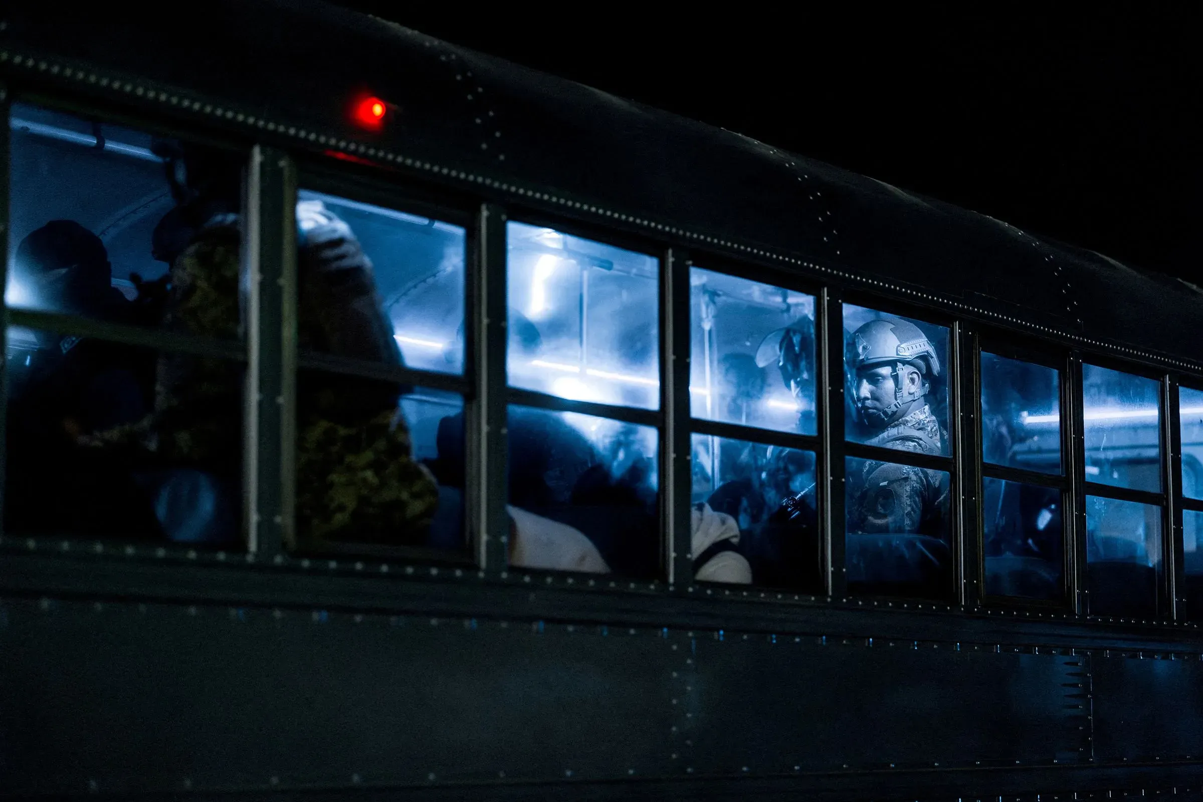 حافلة مظلمة تحمل مهاجرين، مع وجود ضباط يرتدون خوذات، تعكس جهود إدارة ترامب في ترحيل المهاجرين إلى دول ثالثة.