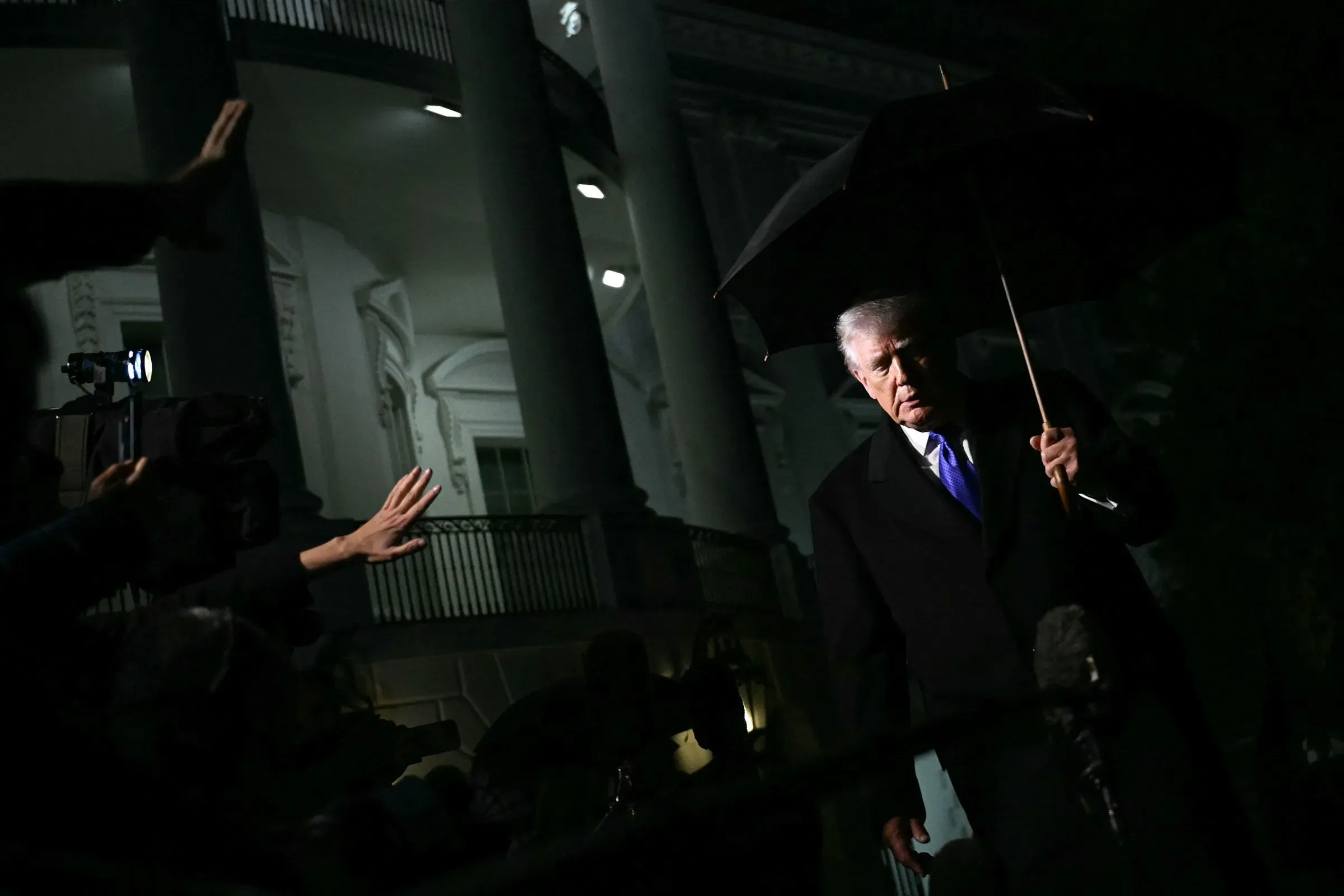 ترامب يحمل مظلة ويتجه نحو الكاميرات في حديقة البيت الأبيض، محاطًا بأضواء الصحفيين، في ظل تصاعد التوترات حول التحقيقات السياسية.