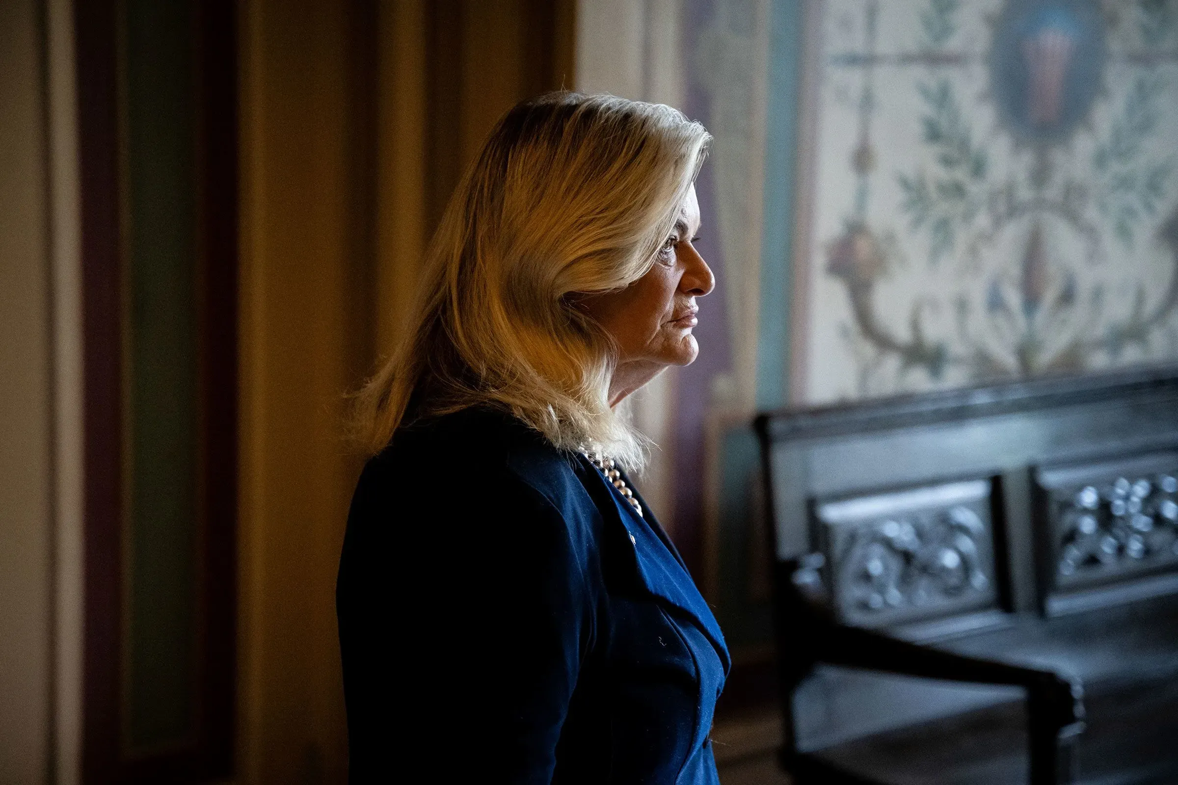 امرأة ذات شعر أشقر طويل ترتدي بدلة زرقاء، تقف في مكان داخلي مضاء بشكل خافت، مع تفاصيل معمارية في الخلفية.