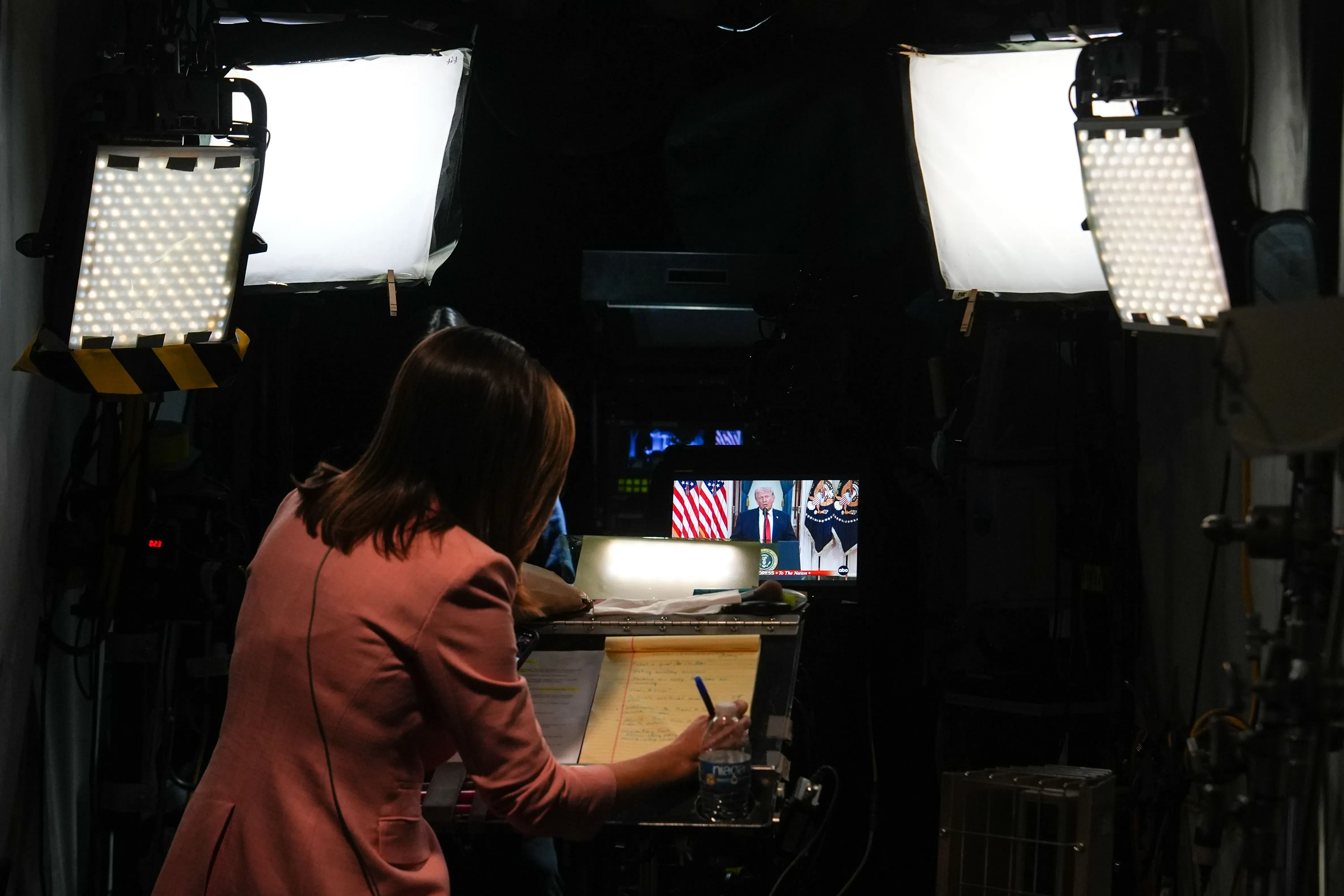 امرأة تعمل على إعداد ملاحظات أثناء بث مباشر لخطاب ترامب في البيت الأبيض، مع ظهور العلم الأمريكي خلفه.