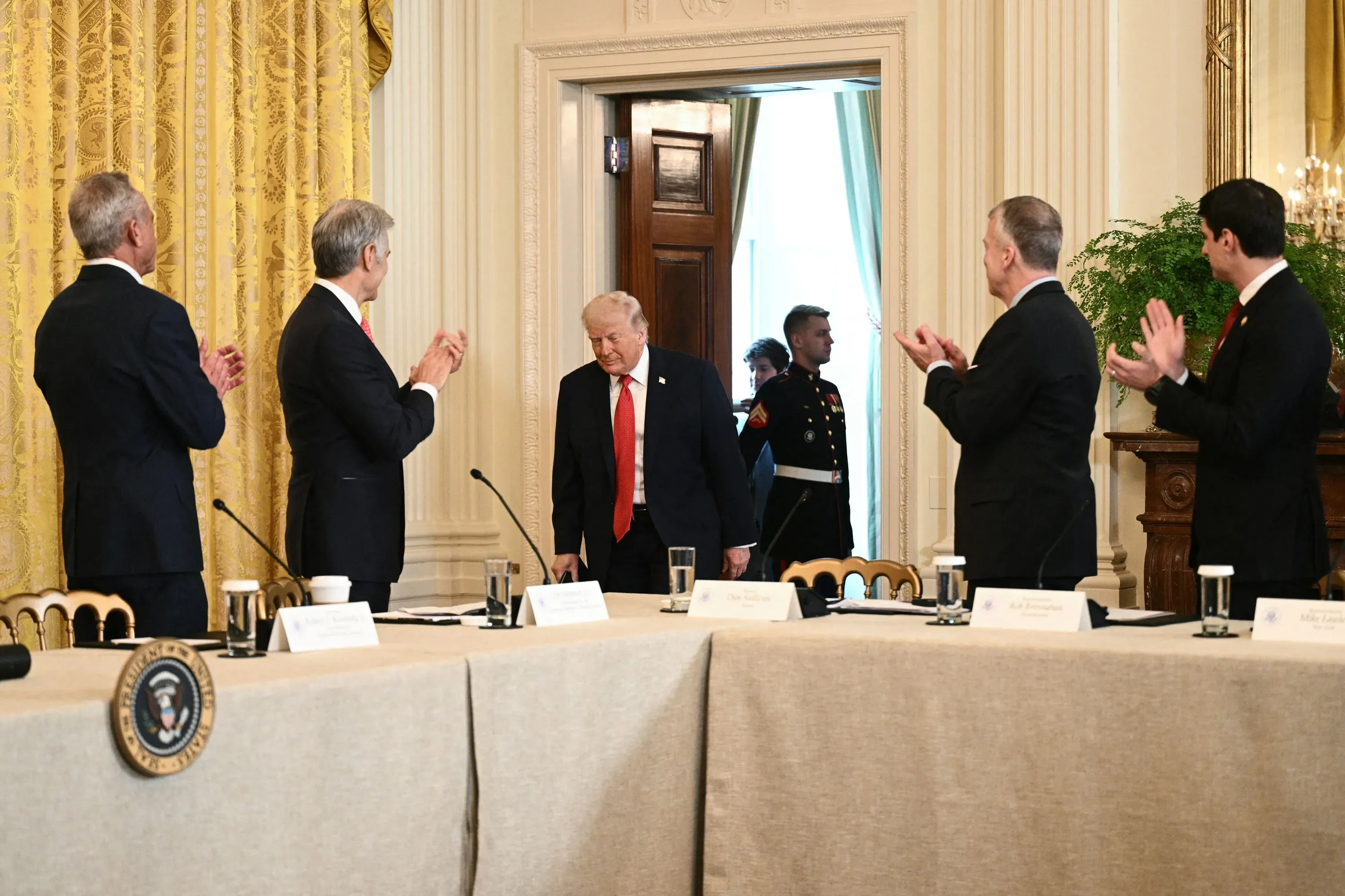 ترامب يدخل قاعة اجتماعات مع مسؤولين، بينما يصفق الحضور. يُظهر المشهد التفاعل الرسمي في سياق السياسة الأمريكية.