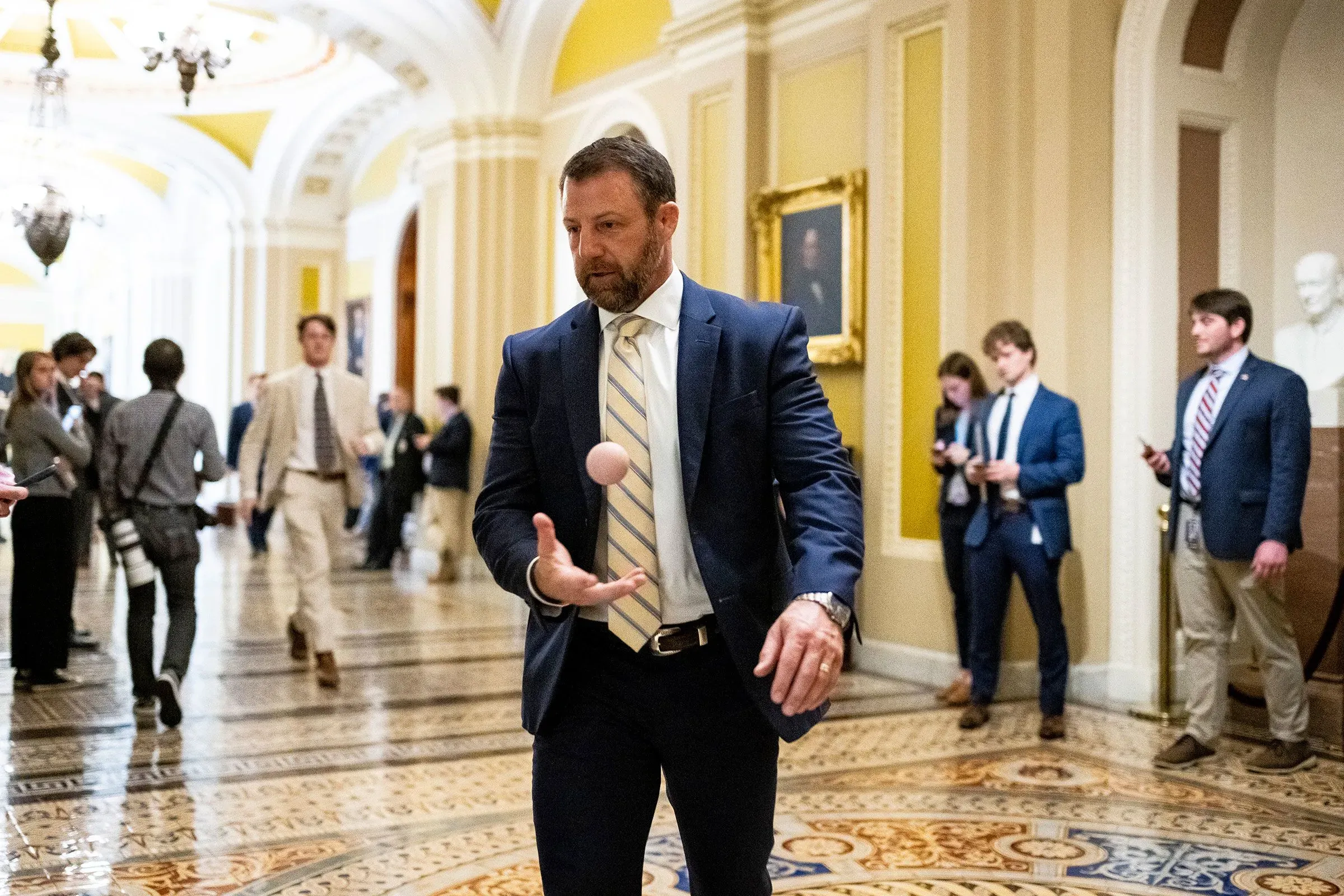 رجل يرتدي بدلة رسمية يقوم بتدوير كرة صغيرة في ممر مبنى حكومي، بينما يظهر عدد من الأشخاص في الخلفية.