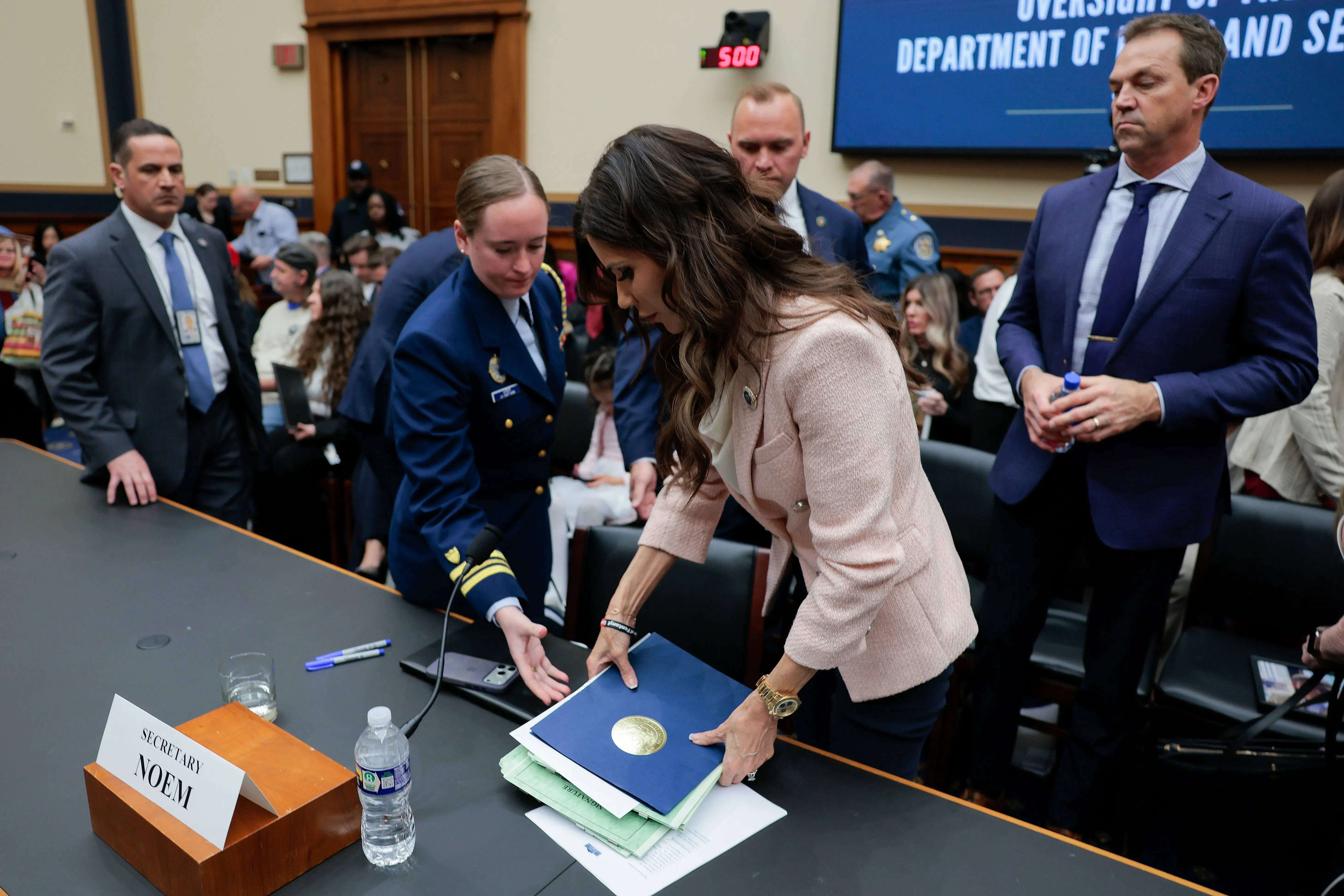 وزيرة الأمن الداخلي كريستي نويم تتحدث في جلسة استماع بالكونغرس، محاطة بمساعدين وموظفين، مع التركيز على الوثائق أمامها.