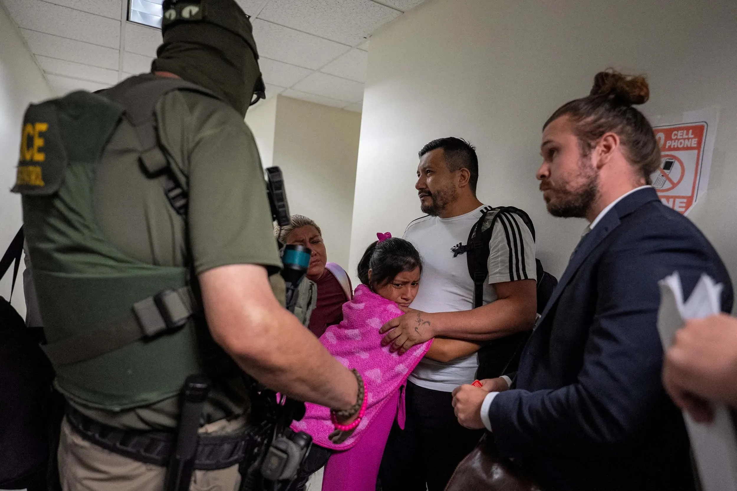 مشهد لعائلة تتعرض للتفتيش من قبل عناصر إنفاذ القانون، مع طفل ملفوف ببطانية وردية، يعكس تأثير سياسات ترامب على المهاجرين.