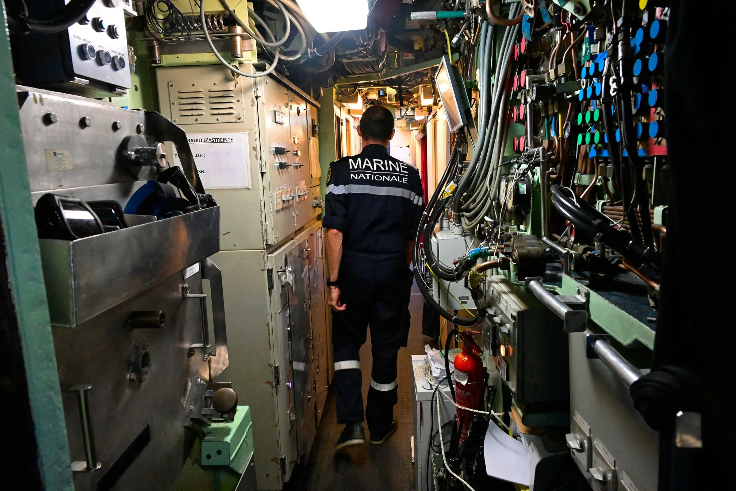 رجل يرتدي زي البحرية الوطنية يسير في ممر ضيق داخل غواصة، محاط بأجهزة ومعدات تقنية، مما يعكس أهمية الدفاع البحري في السياق الجيوسياسي.