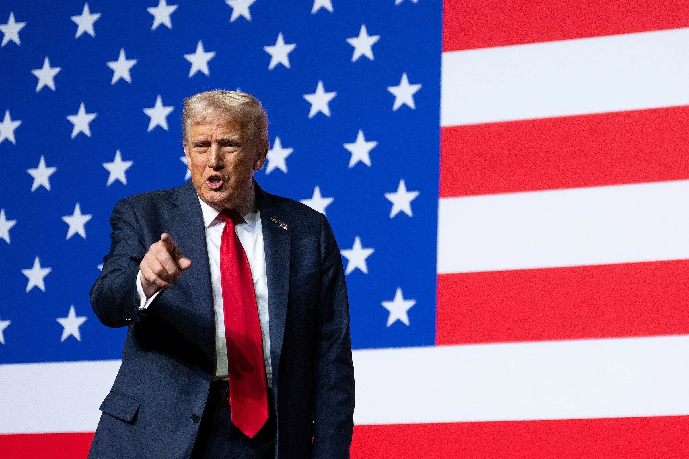 ترامب يتحدث بحماس أمام علم الولايات المتحدة، معبراً عن آرائه حول السياسات الحكومية والتخفيضات المحتملة.