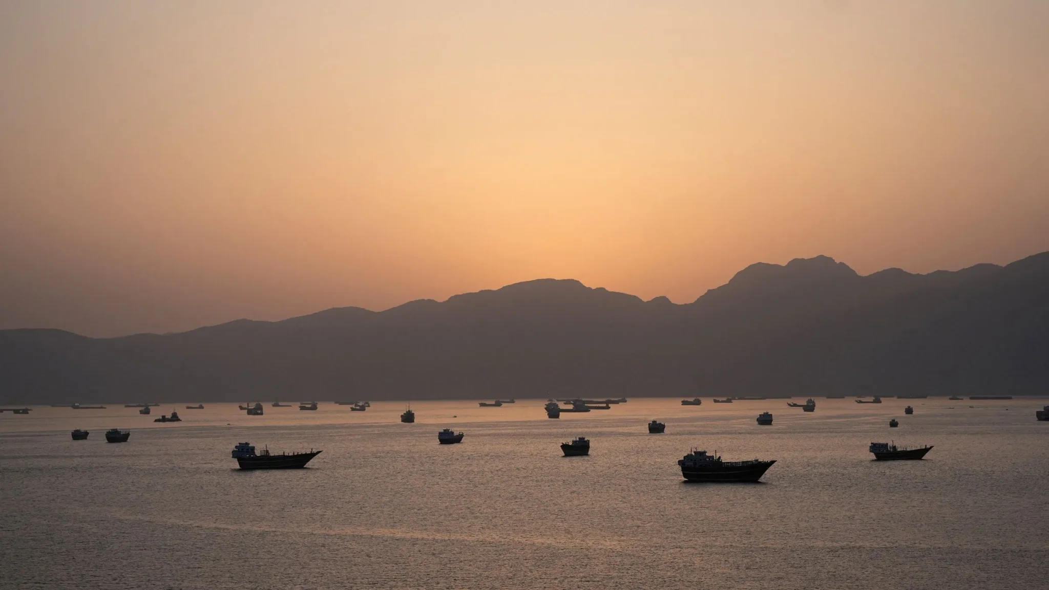 غروب الشمس فوق مضيق هرمز، مع سفن تبحر في المياه الهادئة، يرمز إلى التوترات الجيوسياسية وتأثيرها على المنطقة.