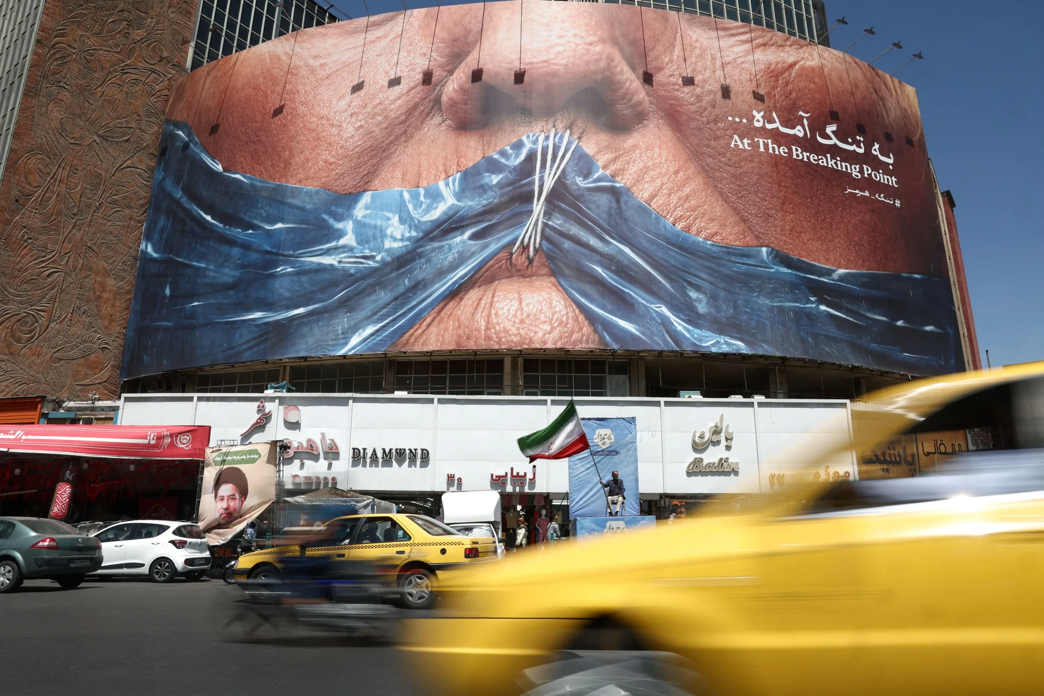 لوحة جدارية ضخمة في طهران تظهر وجهًا مغطى بشريط أزرق مربوط بخيوط، تعبير عن التوتر السياسي بين إيران والولايات المتحدة وتأثيره على الشرق الأوسط.