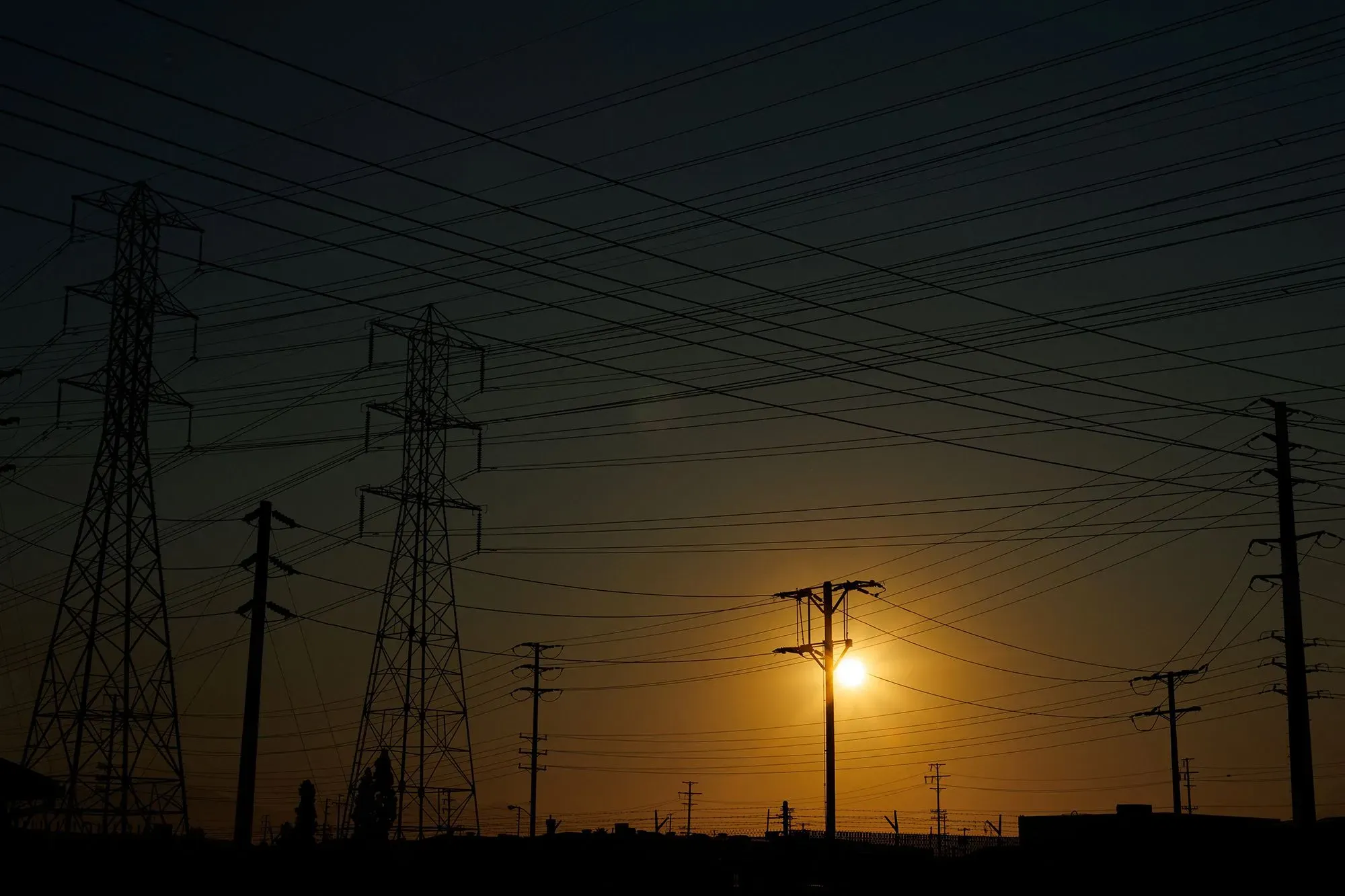 غروب الشمس خلف أعمدة كهرباء، مع تداخل الأسلاك، يعكس التحديات الاقتصادية التي تواجه الأمريكيين في ظل ارتفاع أسعار الوقود.