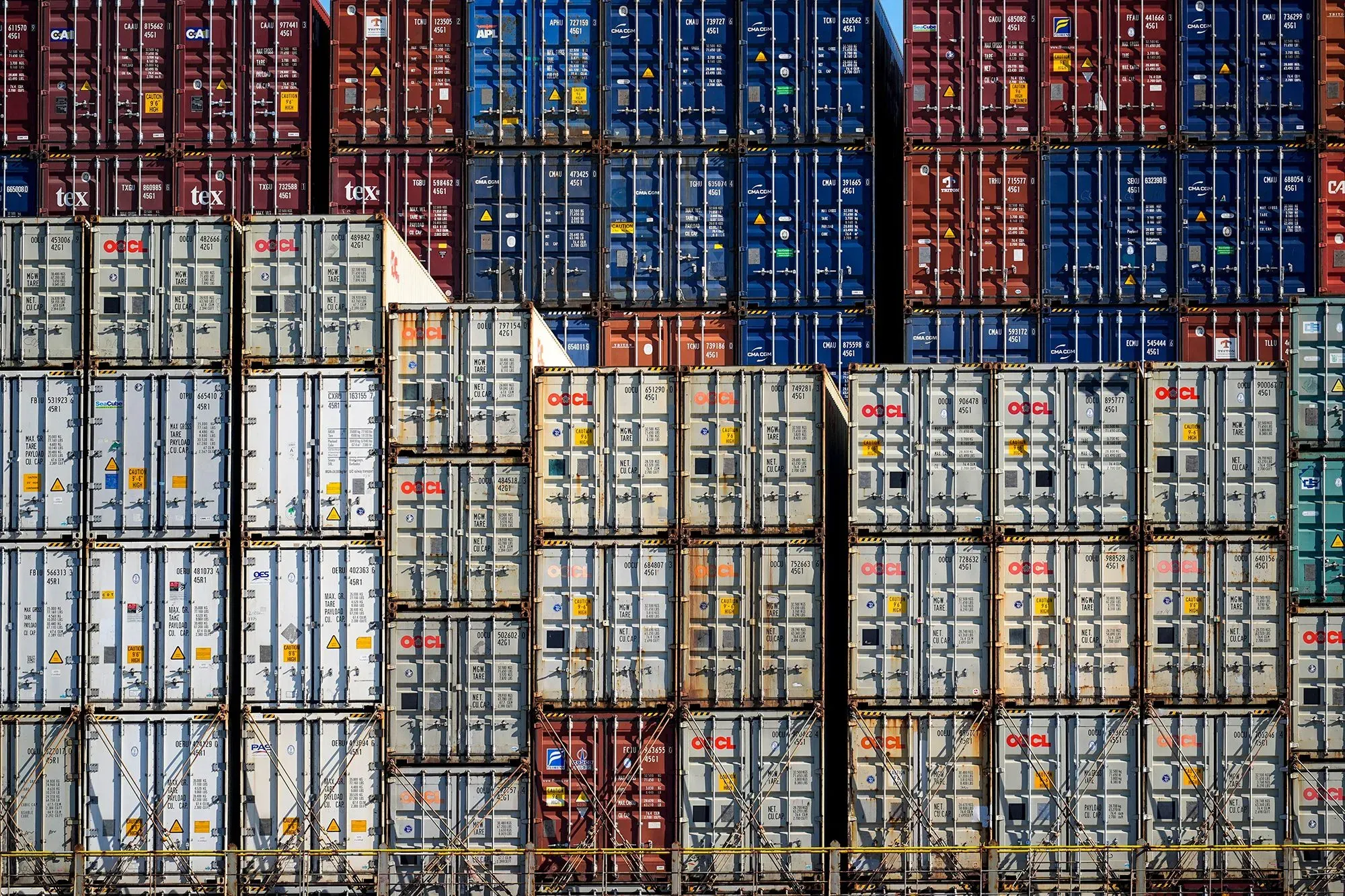 صناديق شحن ملونة مكدسة في ميناء، تعكس تأثيرات سلسلة التوريد على الاقتصاد الأمريكي وتكاليف المستهلك.