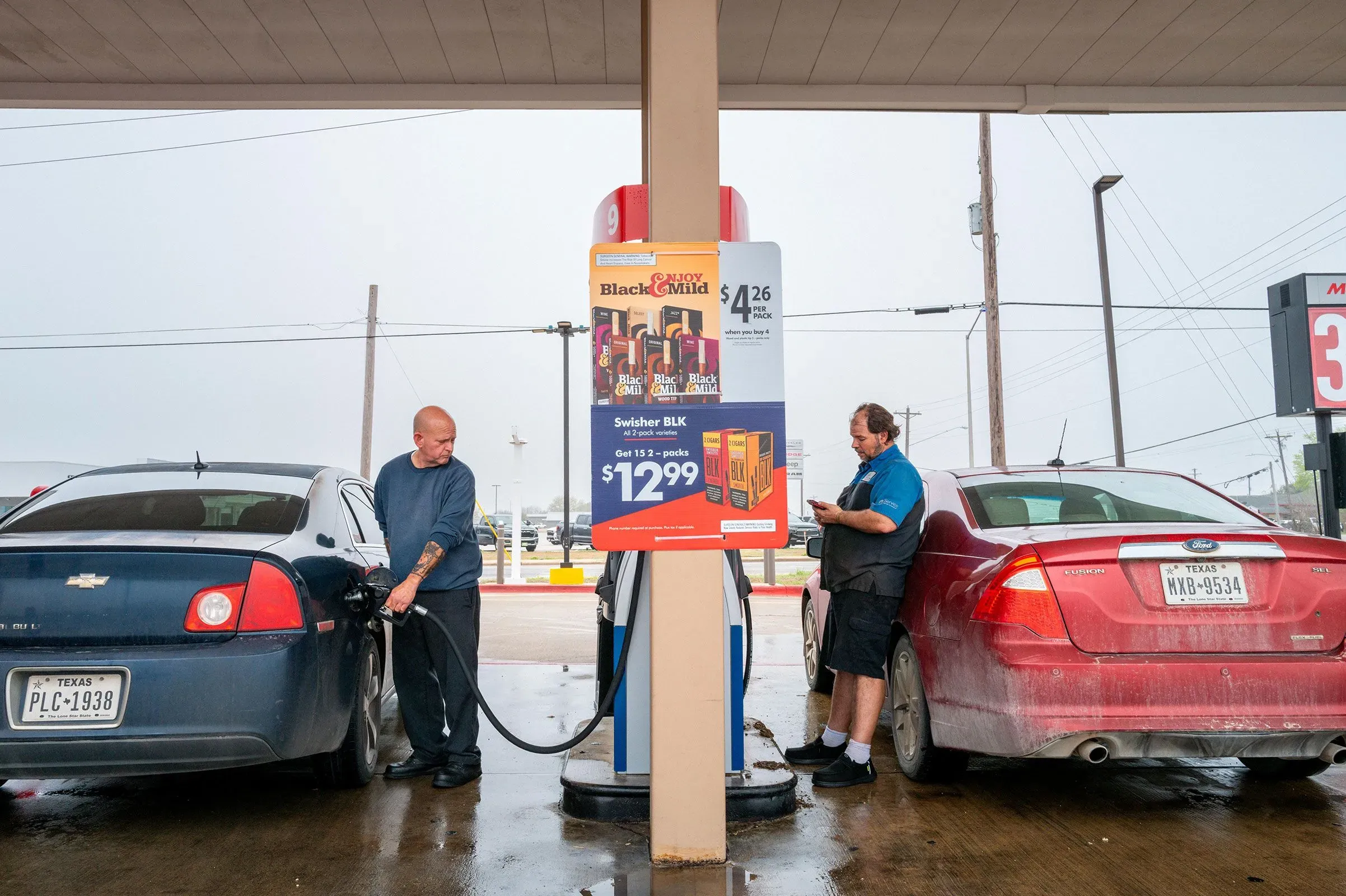 محطة وقود مزدحمة حيث يقوم رجل بتعبئة سيارته بالبنزين، بينما يقف آخر بجوار سيارة أخرى. تظهر لافتة الأسعار في الخلفية.