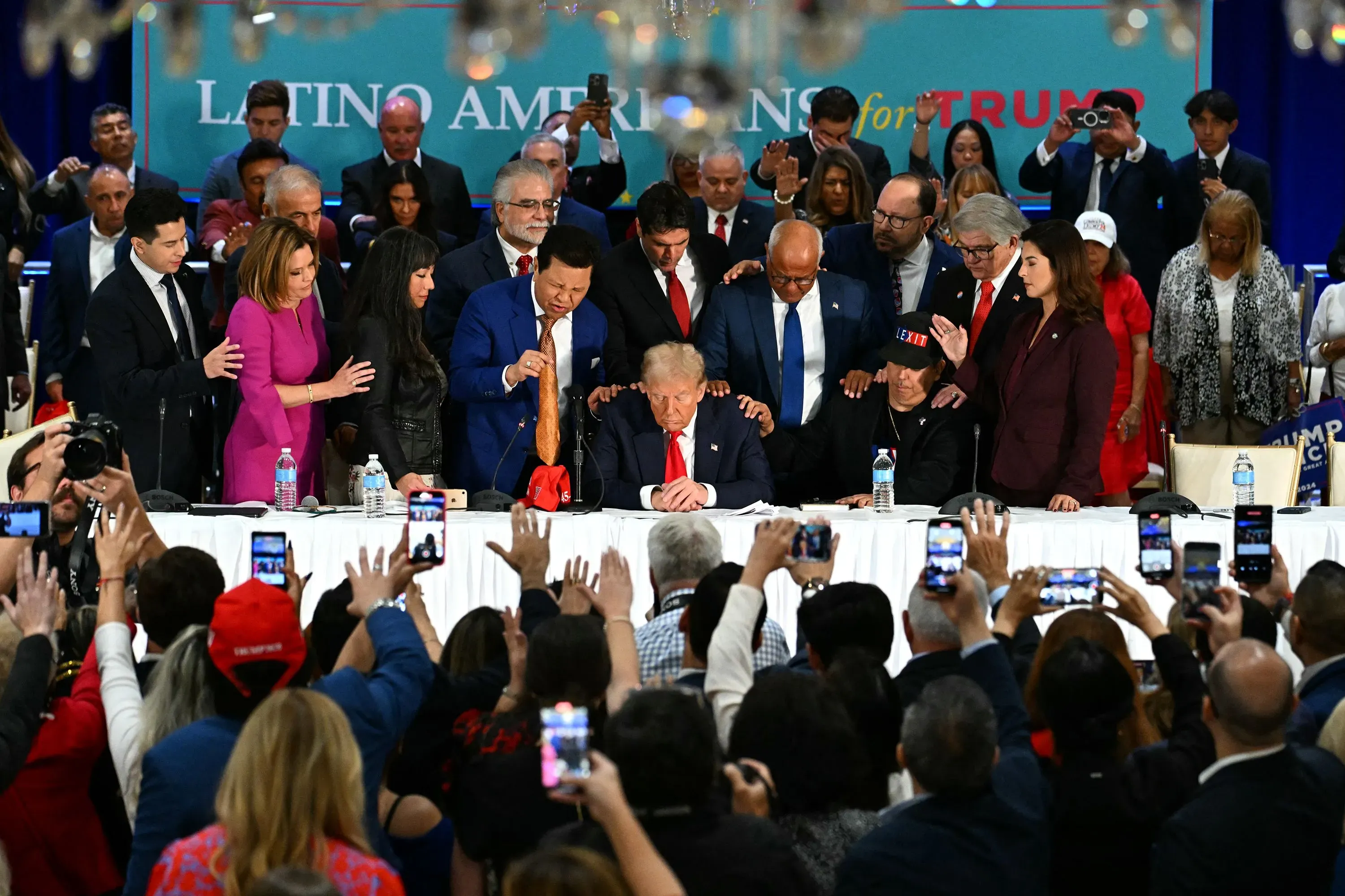 اجتماع لدعم ترامب مع مجموعة من اللاتينيين، حيث يظهر ترامب جالسًا وسط حشد من المؤيدين، مع لافتة "لاتينو أمريكان فور ترامب" في الخلفية.