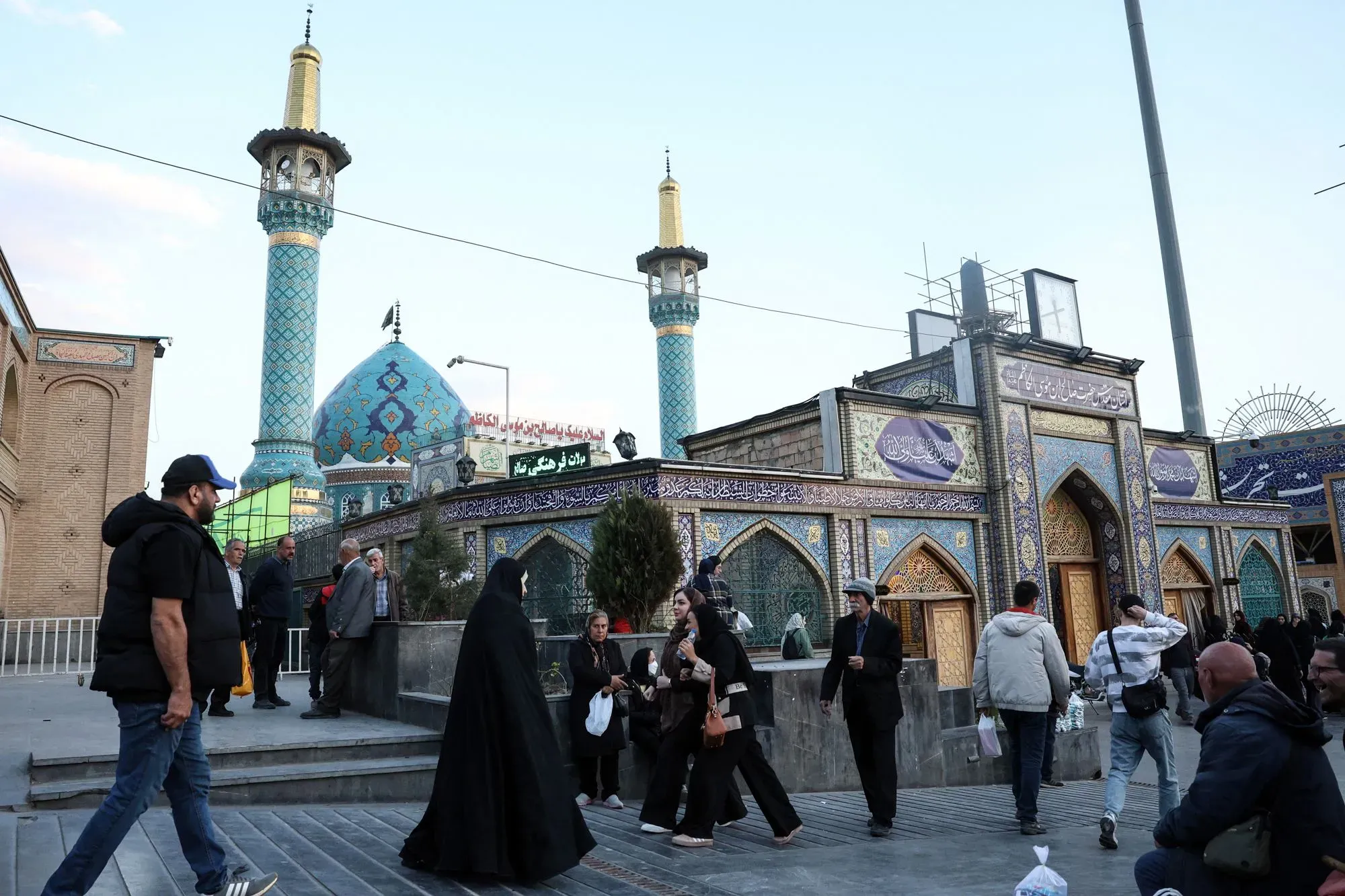 مزار إيراني مزدحم، يظهر فيه أشخاص يمشون بجوار مبانٍ مزخرفة وأبراج مئذنة، مما يعكس الثقافة والدين في المنطقة.