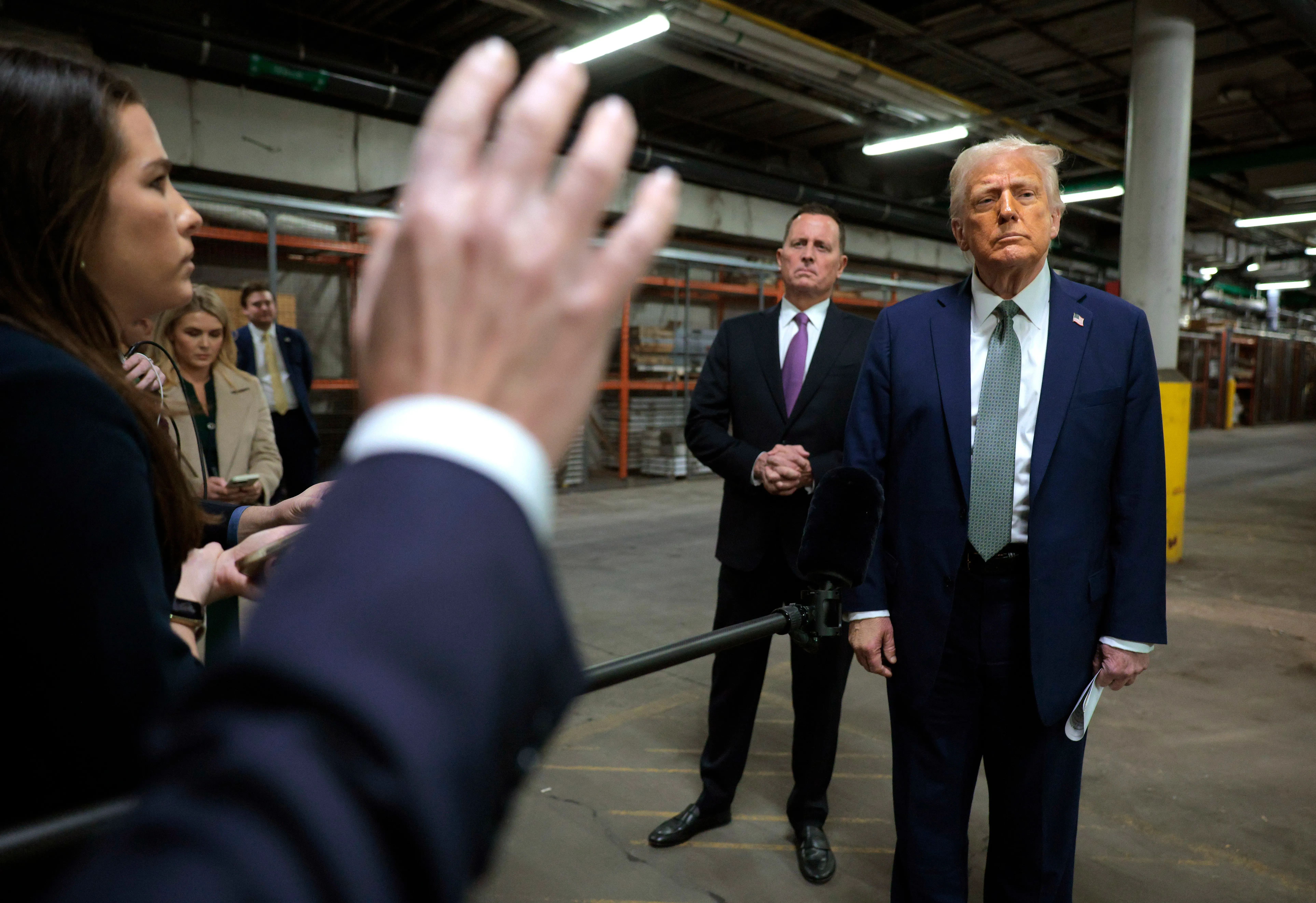 ترامب يتحدث للصحفيين في مركز كينيدي، بينما يقف غرينيل وفلوكا في الخلفية، مما يعكس التوترات حول القيادة والتجديدات.