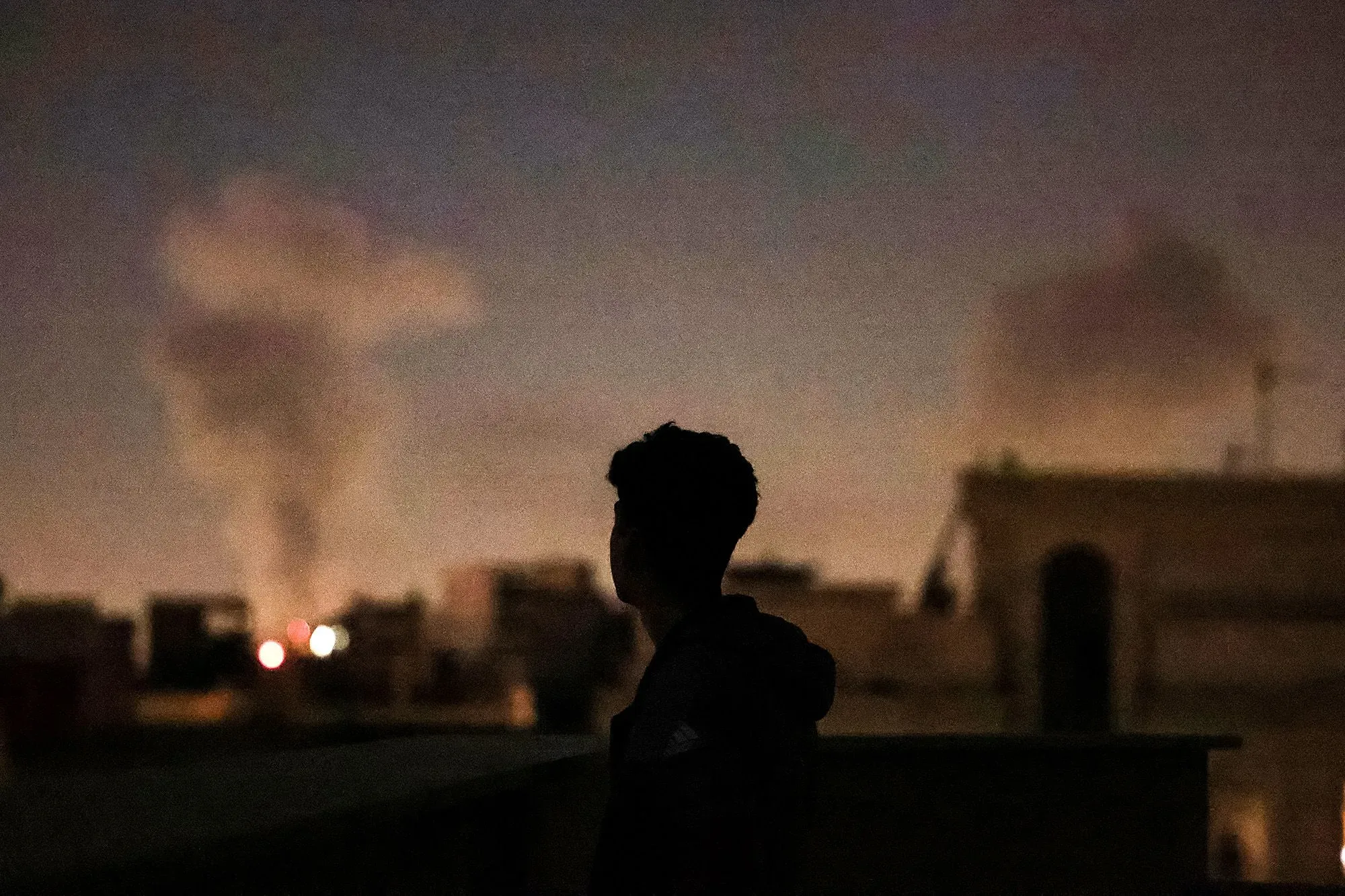 صورة لصبي يقف في مكان مرتفع، يراقب الدخان المتصاعد من انفجارات في الأفق، مما يعكس التوترات في الشرق الأوسط وتأثيرها على الاقتصاد.