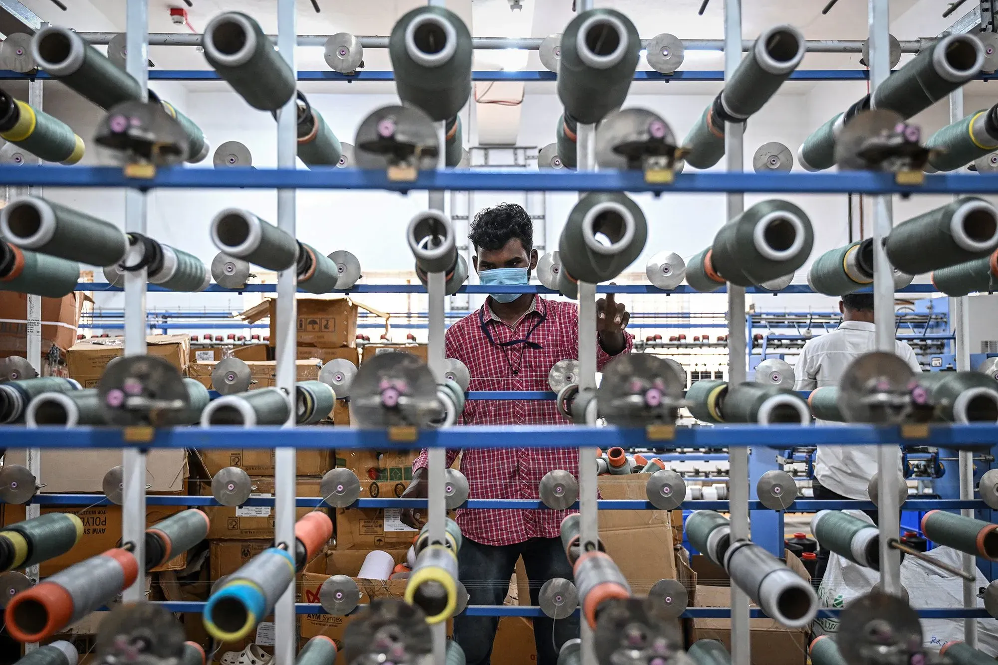 عامل يرتدي كمامة في مصنع، محاط بلفافات من الخيوط الملونة، يعكس أهمية الصناعة النسيجية في الهند.