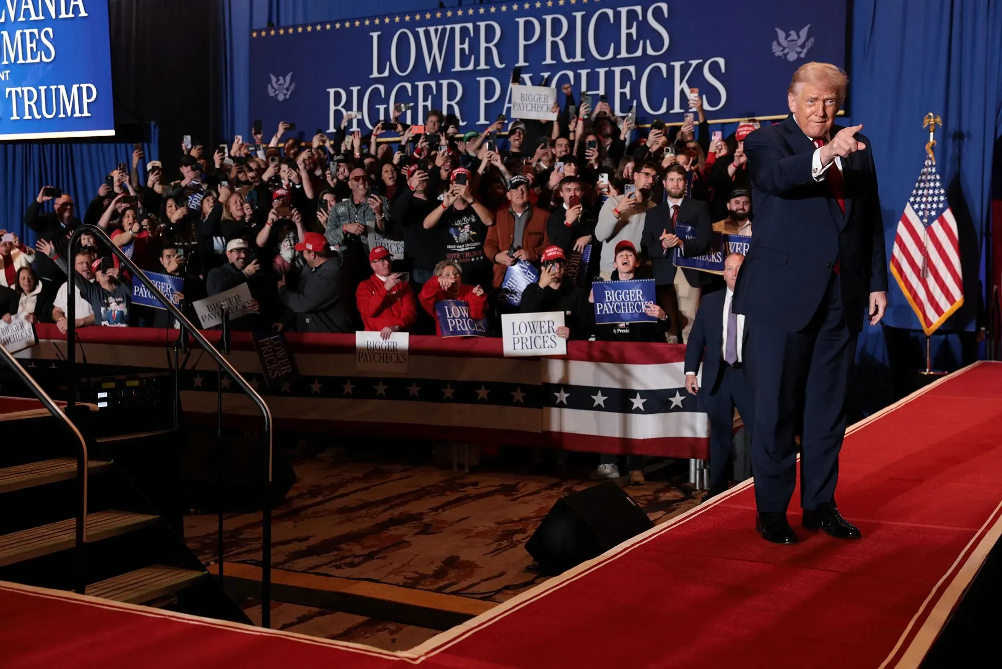 ترامب يتحدث أمام حشد كبير خلال حدث سياسي، مع لافتات تدعو لخفض الأسعار وزيادة الرواتب، مما يعكس قضايا الاقتصاد الحالية.
