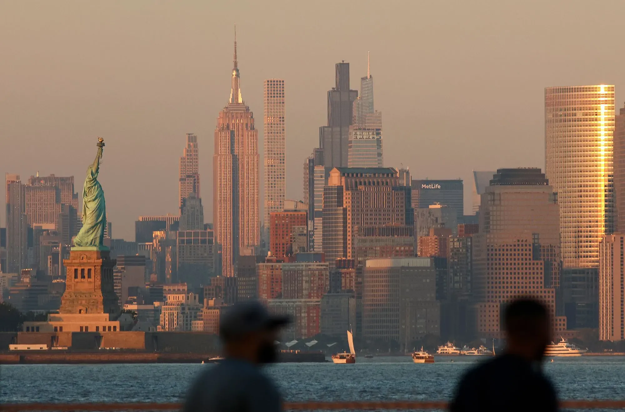 صورة لمدينة نيويورك تظهر تمثال الحرية أمام أفق المدينة المليء بناطحات السحاب في غروب الشمس، مع شخصين في المقدمة بالظل.
