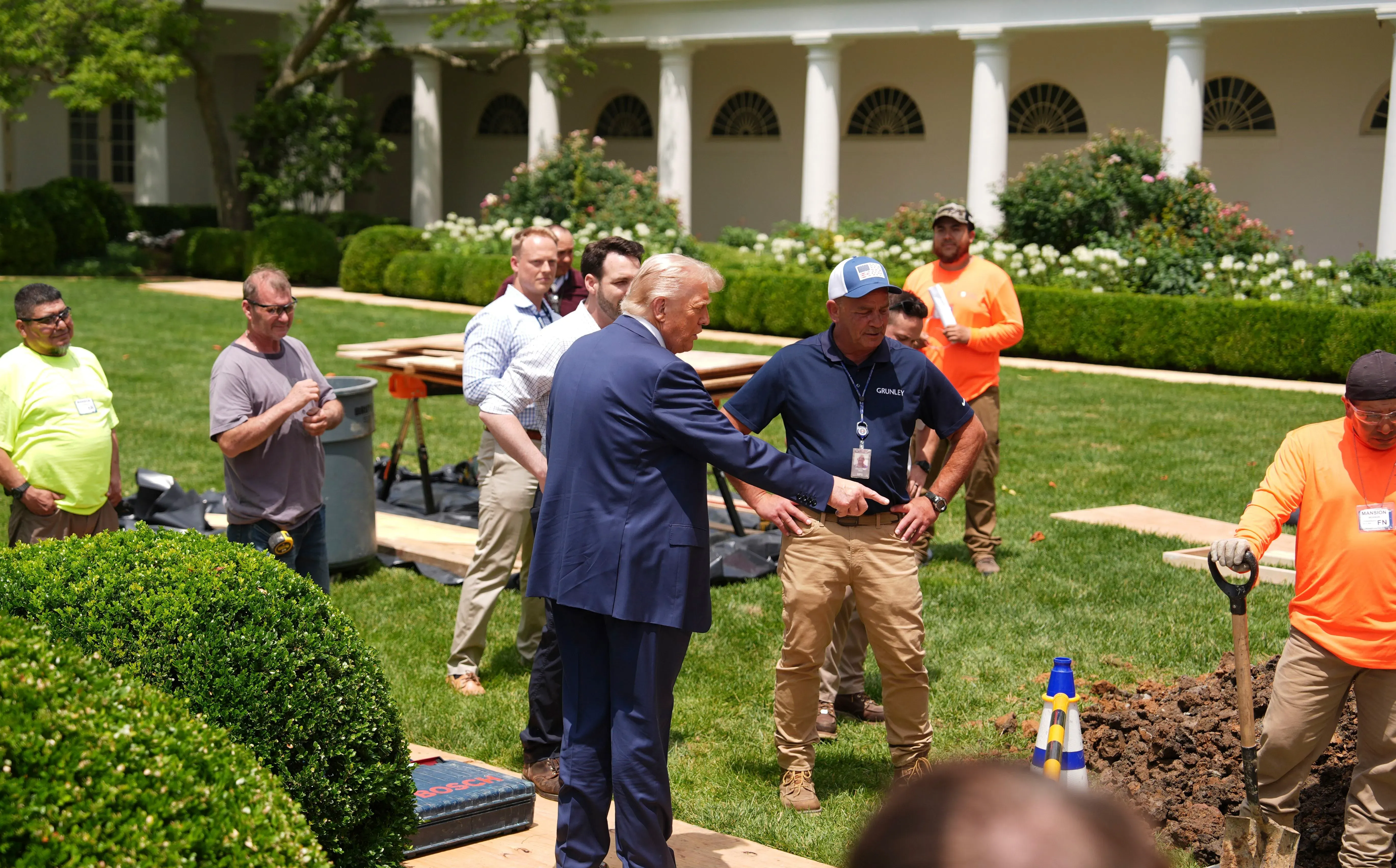 الرئيس دونالد ترامب يتحدث مع مجموعة من العمال في حديقة الورود أثناء تجديدها، مع وجود معدات البناء والأشجار المحيطة.