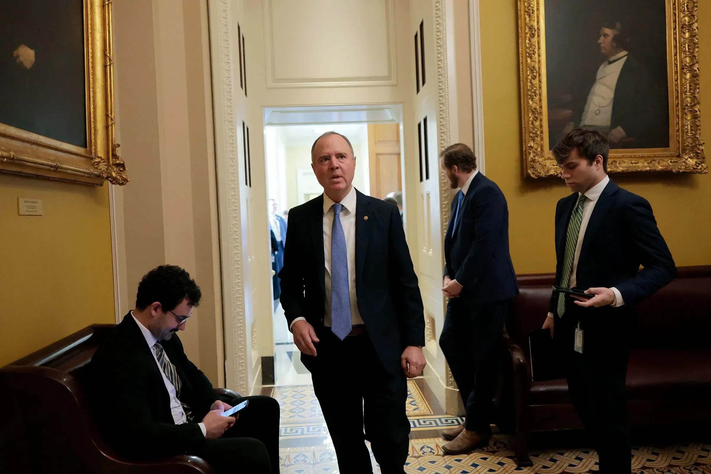 آدم شيف يسير في ممر بمبنى الكابيتول، محاطًا بمساعديه، مع لوحات فنية تاريخية على الجدران، في سياق تحقيقات سياسية معقدة.