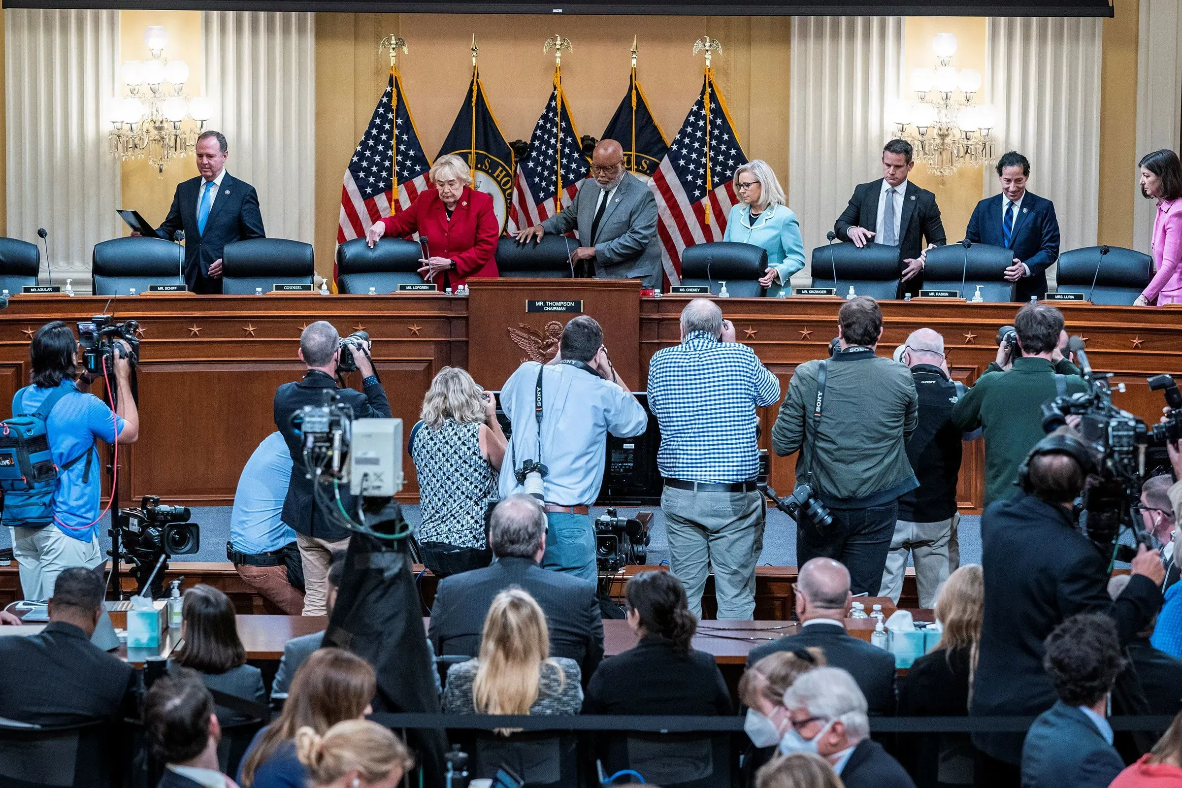 اجتماع لجنة مجلس النواب مع أعضاء يتجهزون للمناقشة، بينما يلتقط المصورون الصور. تظهر الأعلام الأمريكية خلفهم، مما يبرز أهمية الحدث.