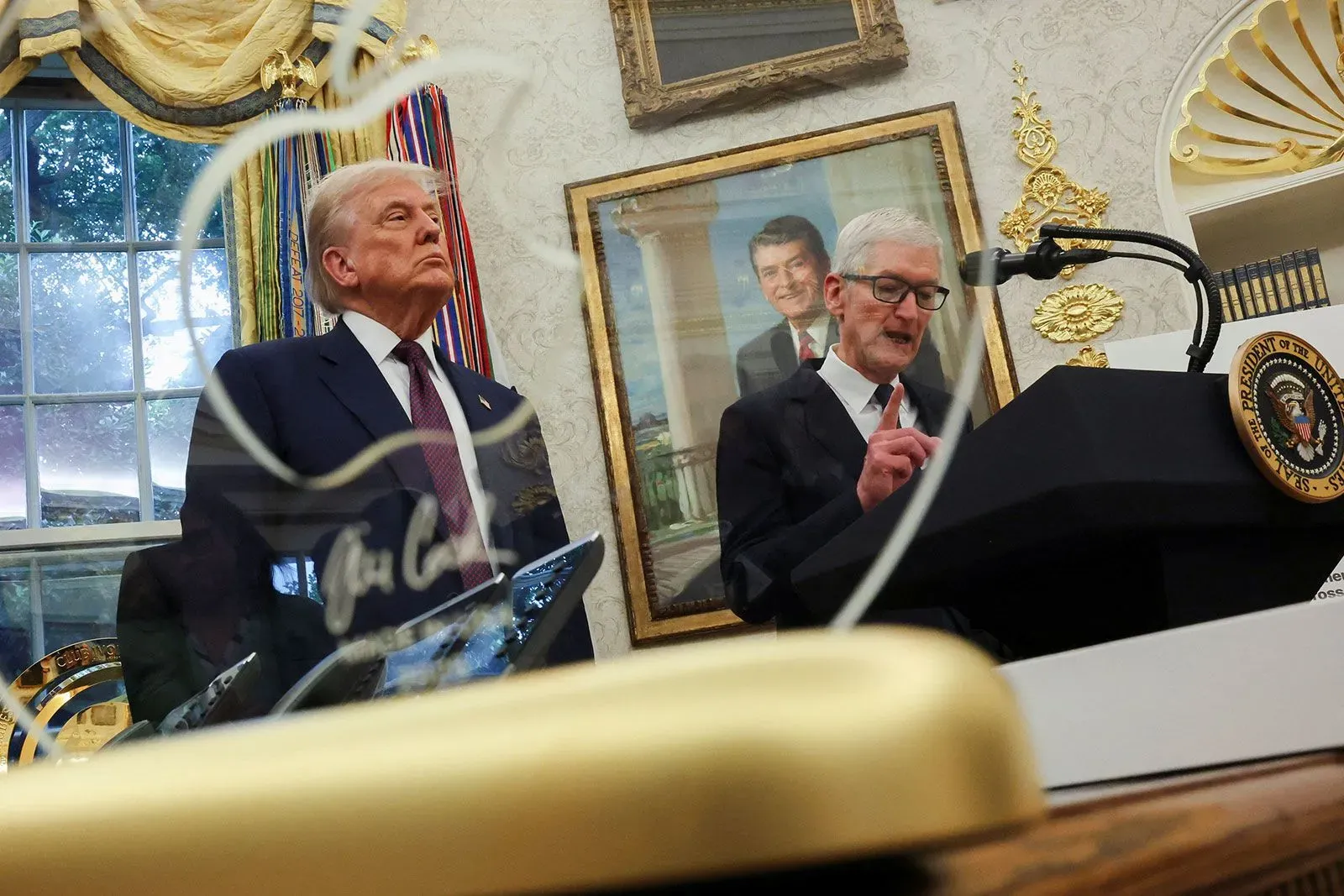 ترامب يستمع إلى تيم كوك، الرئيس التنفيذي لشركة آبل، خلال حدث في المكتب البيضاوي، مع صور تاريخية في الخلفية.