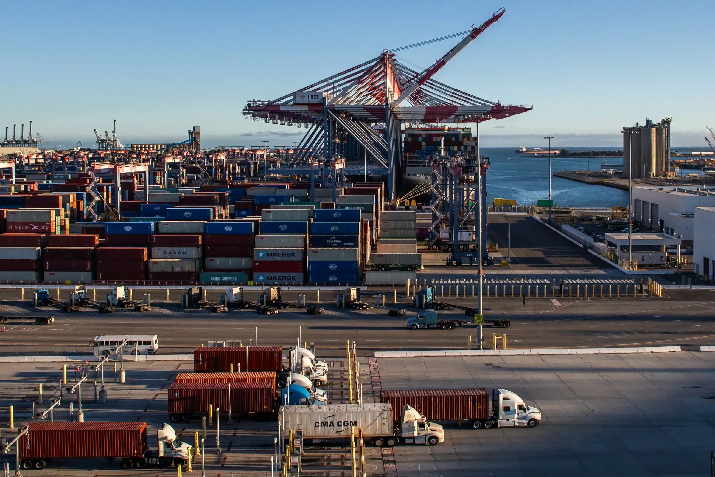 صورة لميناء مزدحم يحتوي على حاويات شحن ملونة وشاحنات نقل، مما يعكس تأثير التعريفات الجمركية على الاقتصاد الأمريكي.