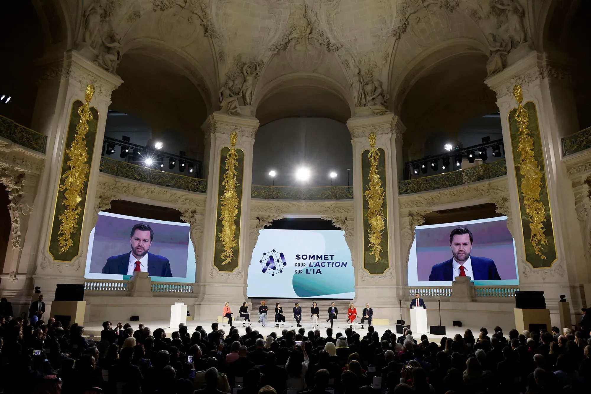 مؤتمر حول الذكاء الاصطناعي في قاعة فخمة، حيث يتحدث المتحدثون أمام جمهور كبير، مع عرض تقديمي خلفهم.