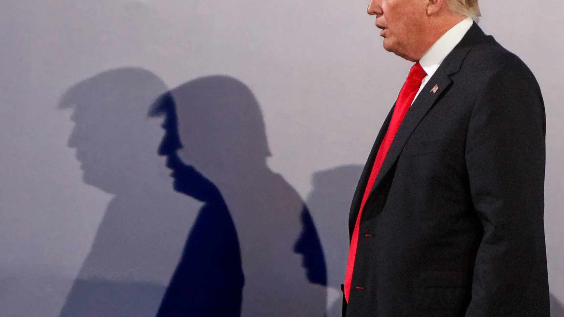 صورة لدونالد ترامب يقف بجانب جدار، حيث تظهر ظلاله بشكل بارز. تعكس الصورة أجواء التوتر السياسي الذي يحيط بممارساته.