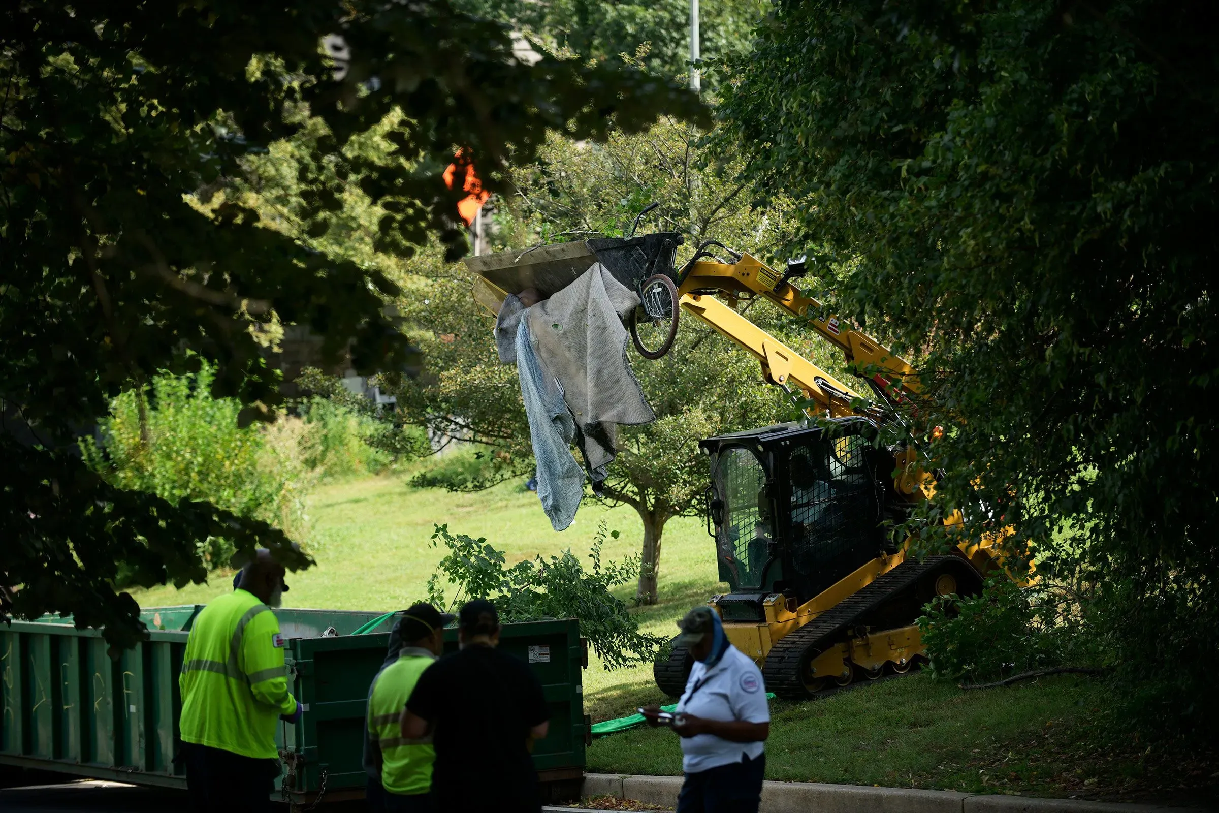 عملية إزالة مخيمات المشردين في منطقة شجرية، حيث يتم رفع الأغراض بواسطة آلية، مع وجود عمال في الخلفية.