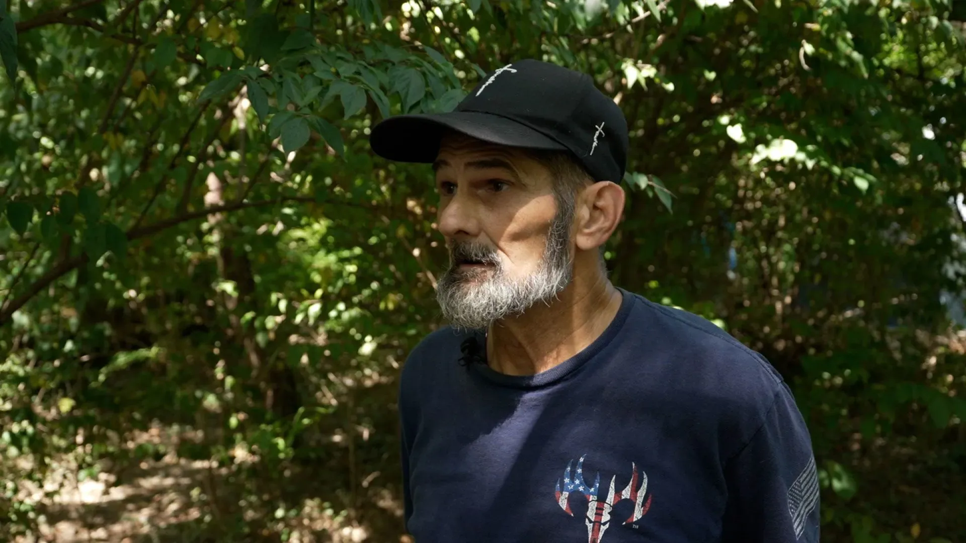 رجل بلا مأوى يتحدث في غابة بفيرجينيا، محاط بالأشجار، مع تعبير وجهه يعكس القلق والتحديات التي يواجهها.