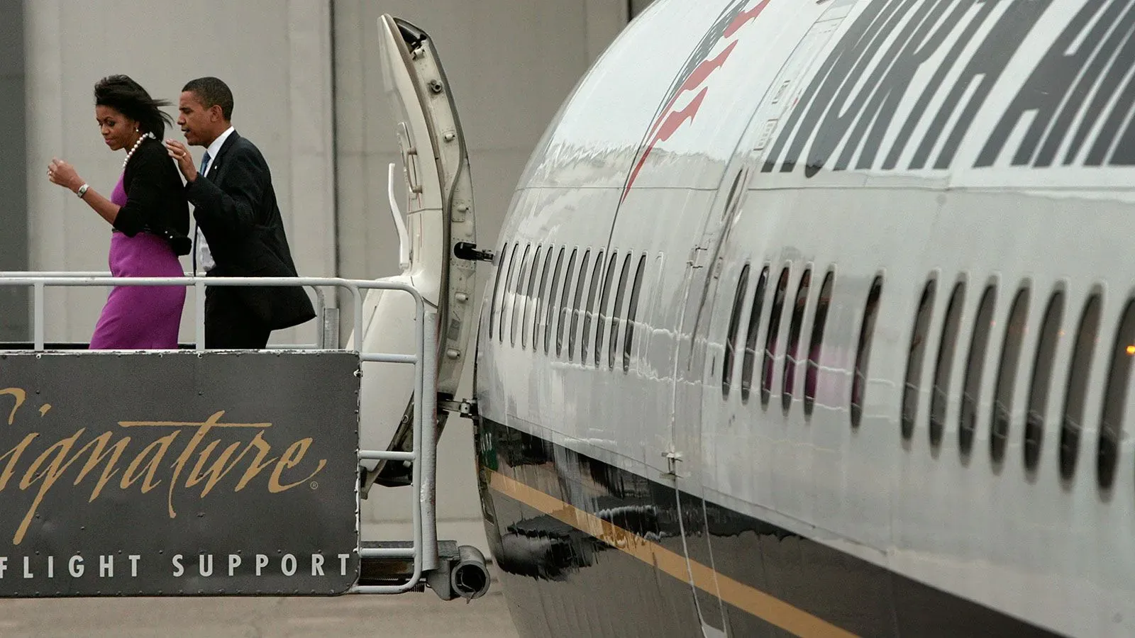 باراك أوباما وميلانيا أوباما يصعدان إلى الطائرة، مع لافتة تشير إلى دعم الرحلات، بينما تظهر الطائرة بلونها الأسود والأبيض.