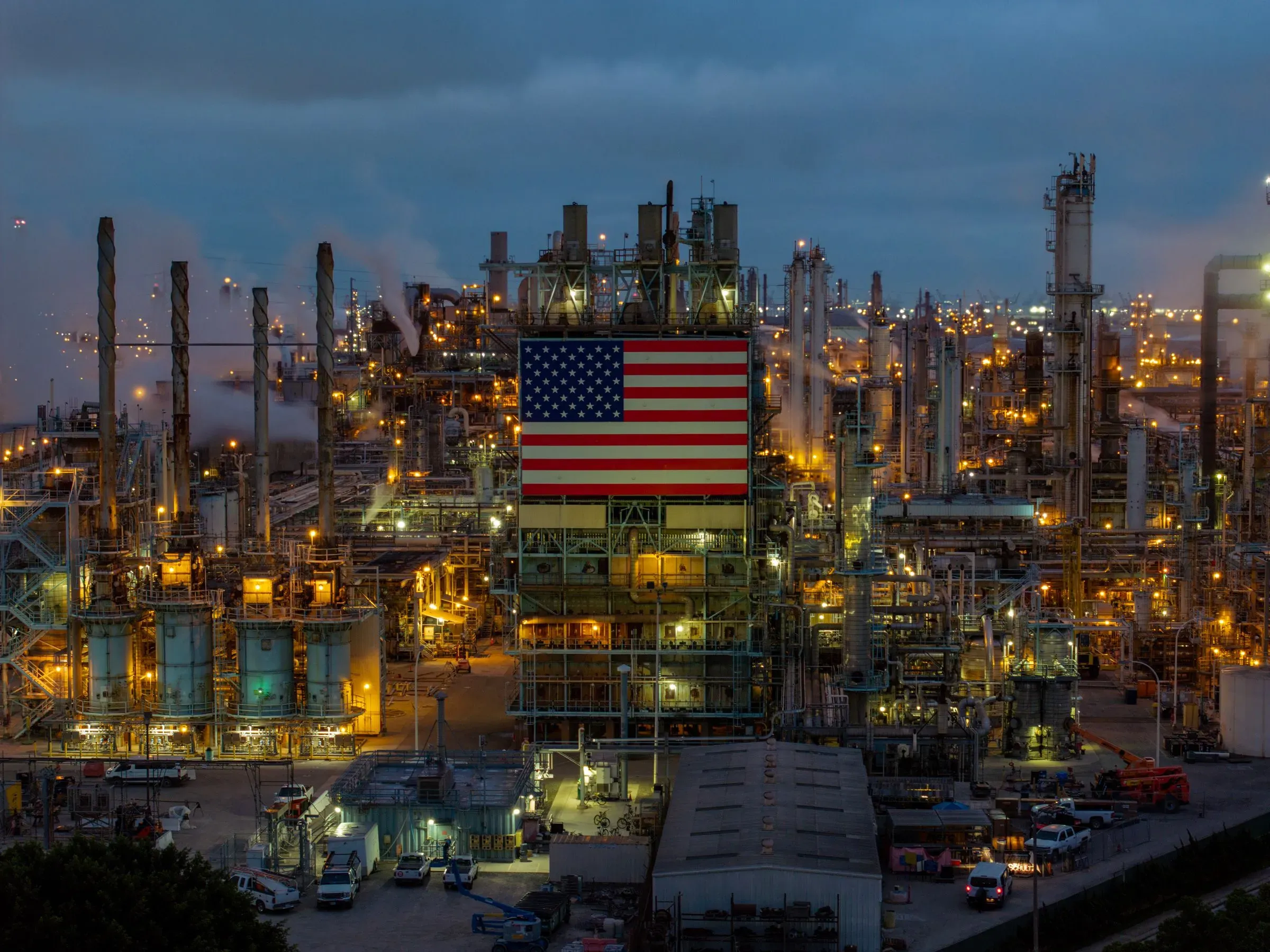 مصنع نفط أمريكي مضاء ليلاً، مع علم الولايات المتحدة كبير على أحد المباني، يعكس أهمية الصناعة النفطية في الاقتصاد الأمريكي.