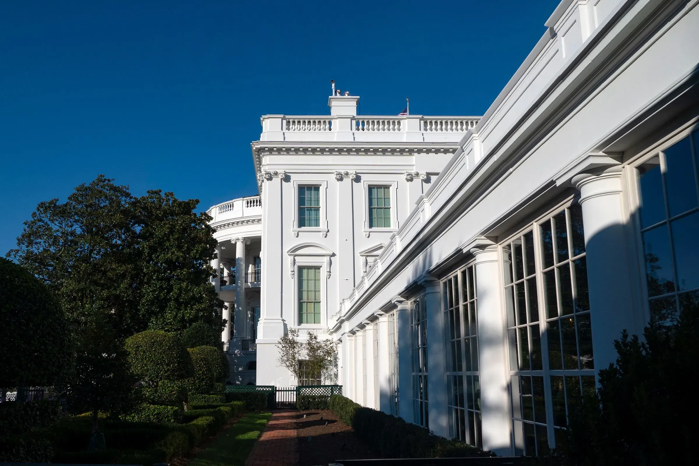 واجهة البيت الأبيض تظهر تفاصيل العمارة الكلاسيكية، مع أشجار وحدائق محيطة، في إطار زمني مشمس.