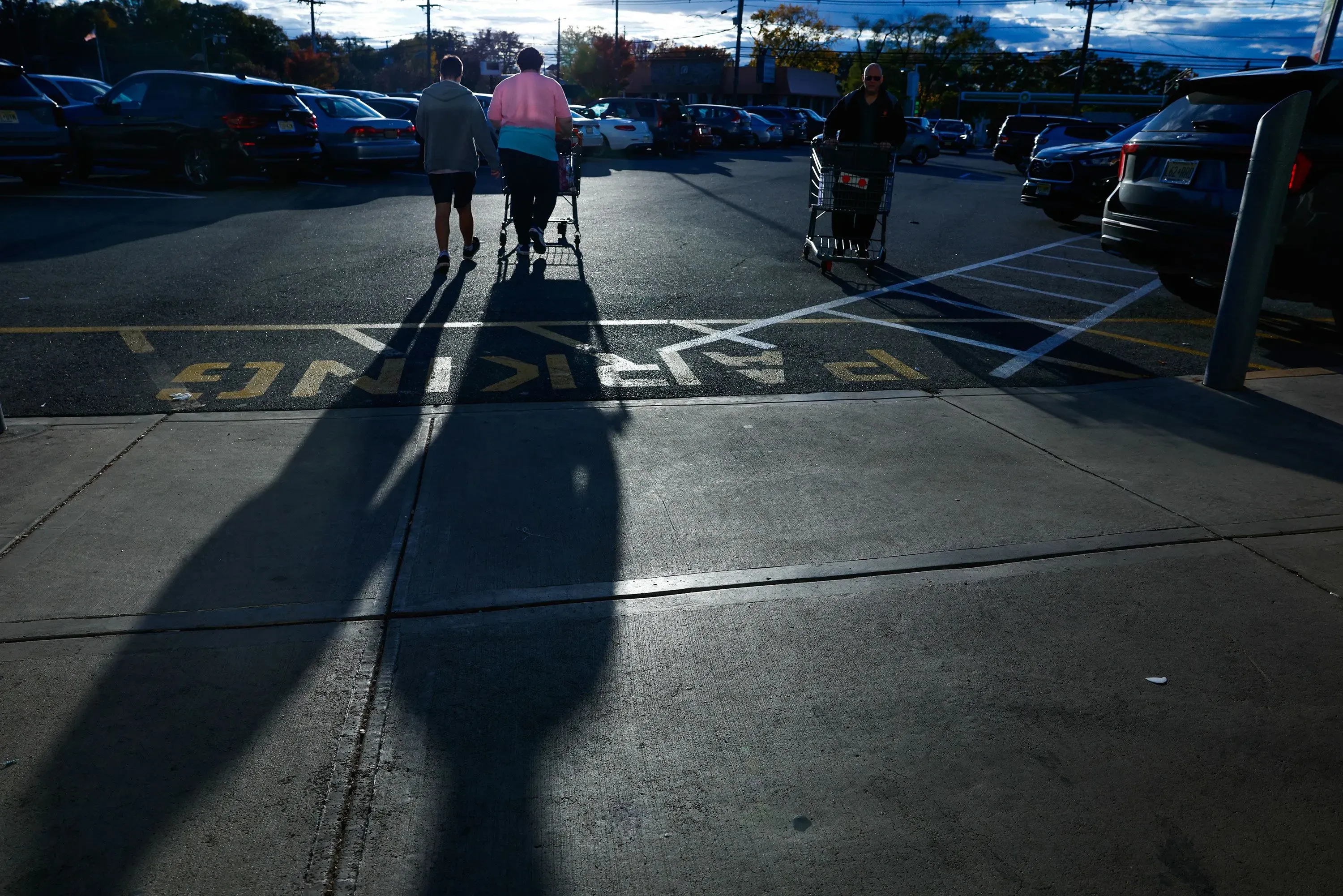 مجموعة من الأشخاص يسيرون في موقف سيارات، مع وجود ظلالهم الطويلة على الأرض، مما يعكس انشغالهم بالتسوق في ظل ارتفاع تكاليف المعيشة.