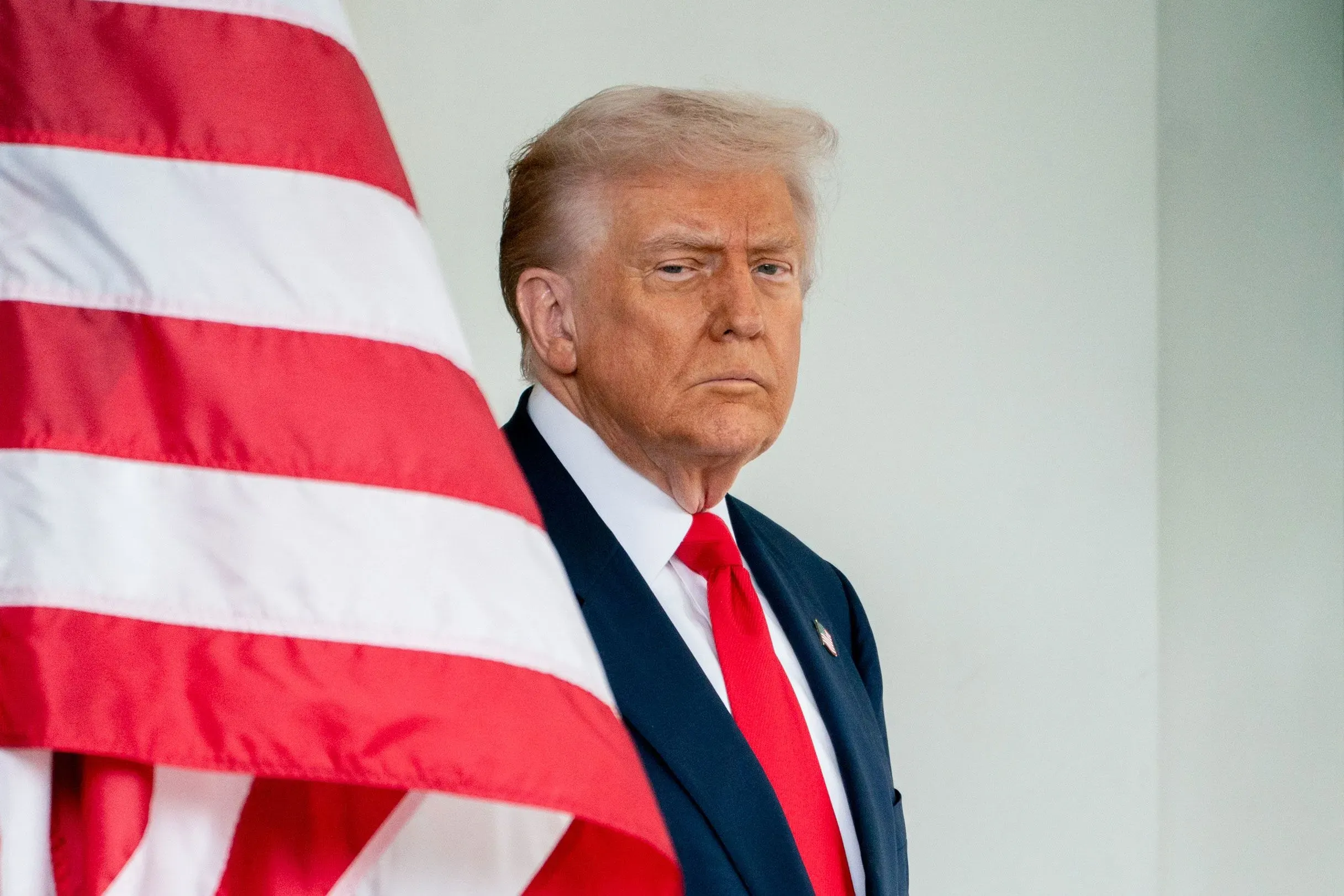 صورة لدونالد ترامب يقف بجانب علم الولايات المتحدة، مع تعبير جاد، مما يعكس توتر الأوضاع السياسية والاجتماعية في البلاد.