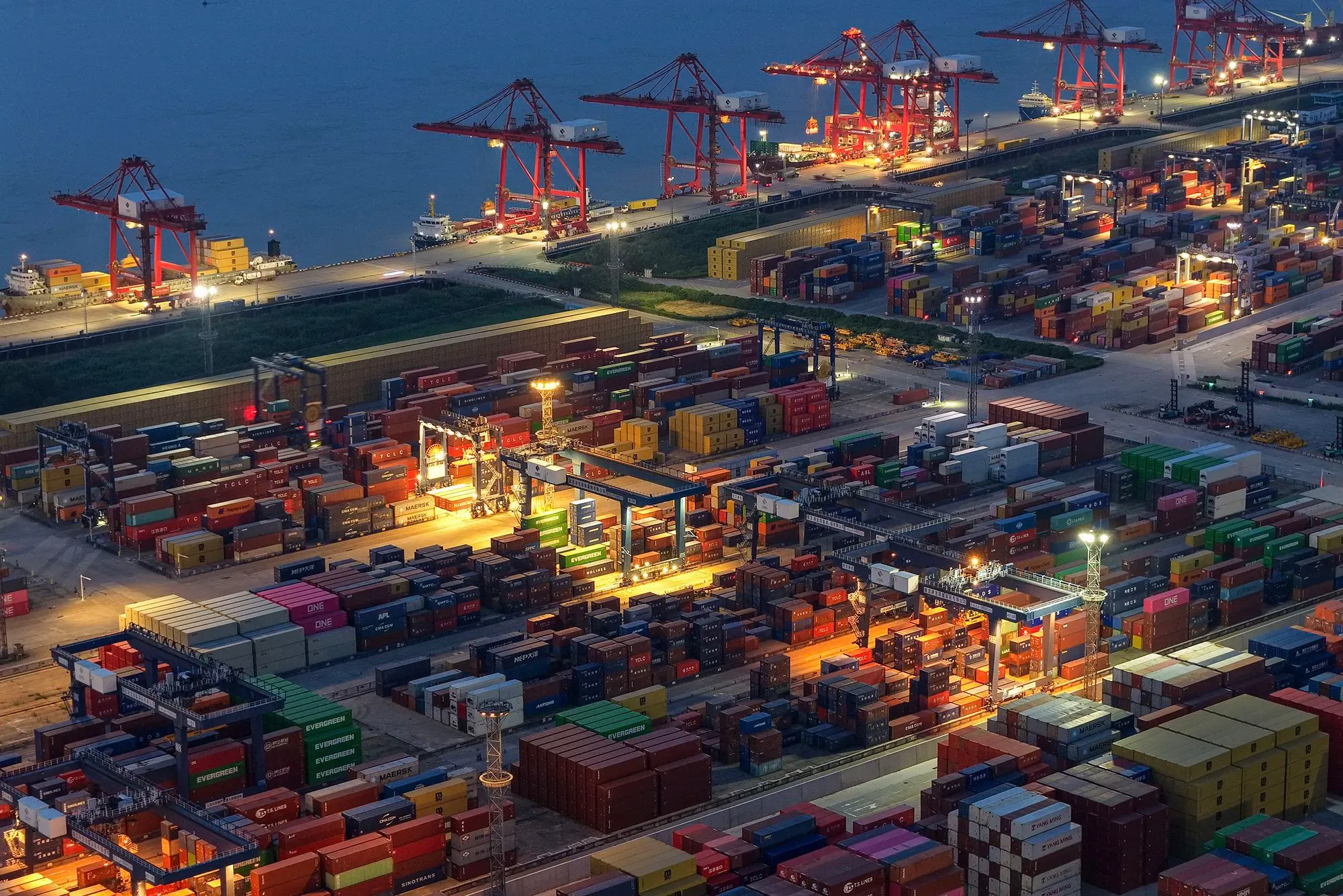 صورة لميناء مزدحم في الليل، حيث تُظهر حاويات شحن ملونة وآلات تحميل، مما يعكس النشاط التجاري وتأثير الرسوم الجمركية على الواردات.