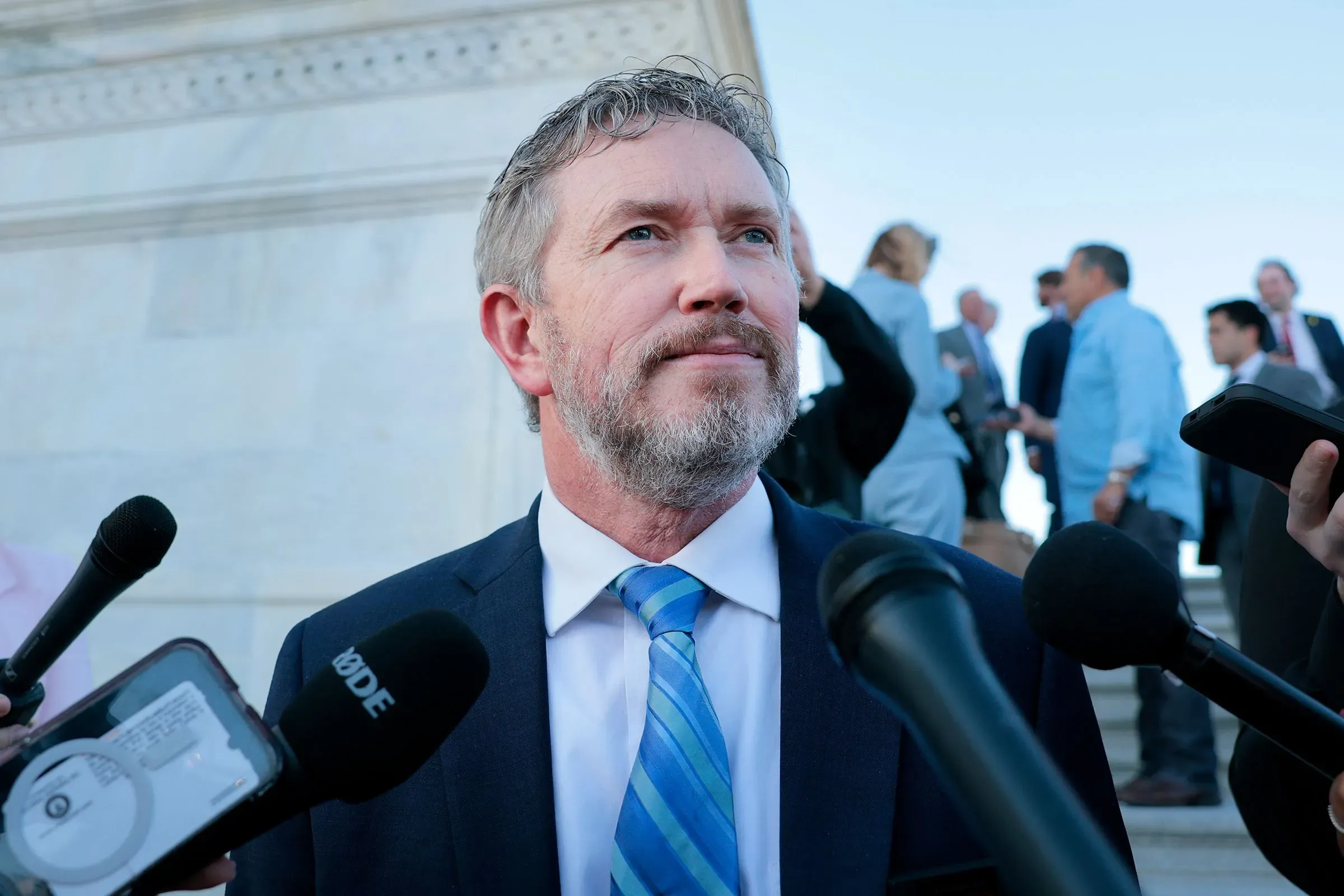 رجل يتحدث إلى الصحافة خارج مبنى حكومي، محاطًا بعدد من المراسلين، في سياق جمع التبرعات لدعم ترامب.