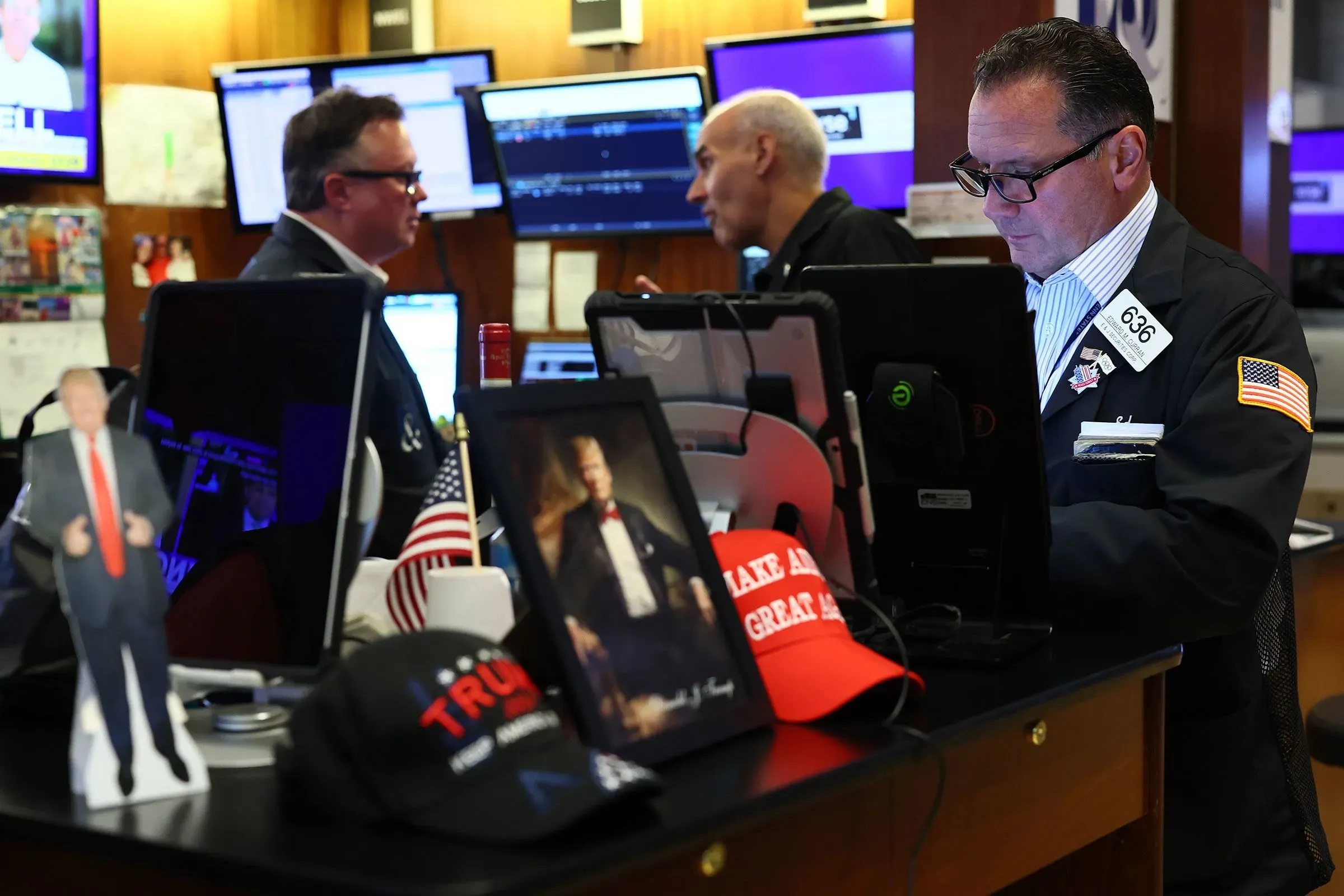 صورة لمتداولين في بورصة، يظهر فيها تمثال صغير لترامب وصورة له، مع قبعة تحمل شعار "اجعل أمريكا عظيمة مرة أخرى"، تعكس أجواء التجارة والتعريفات الجمركية.