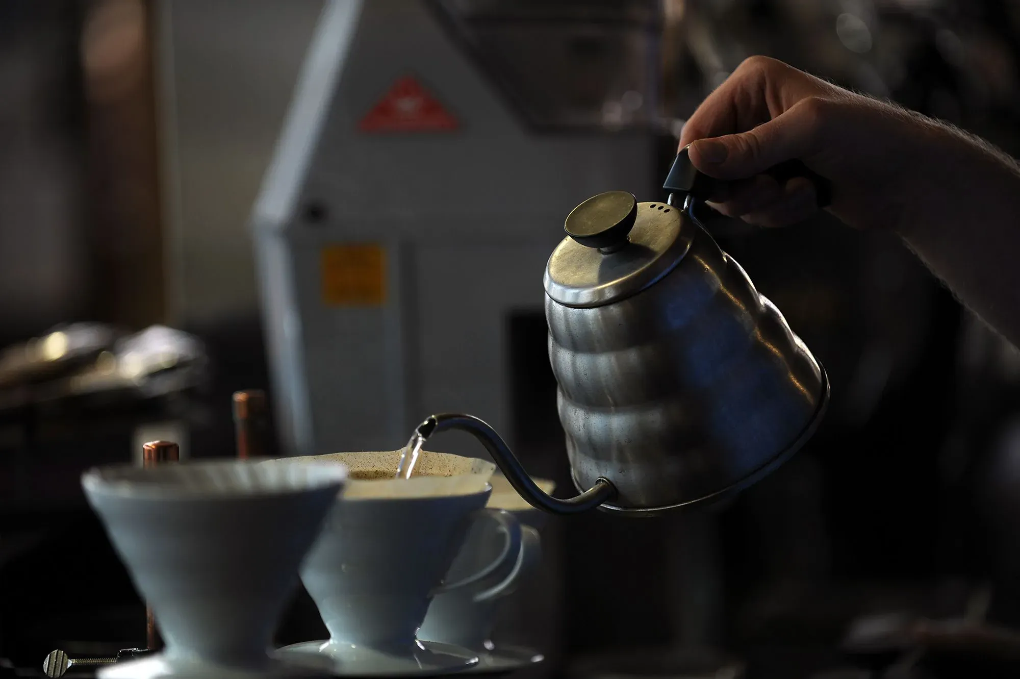 يد لتمسك بإبريق معدني يصب الماء الساخن في فنجان قهوة، مع وجود أدوات تحضير القهوة في الخلفية. تعكس الصورة تزايد أسعار القهوة وتأثيرها على المستهلكين.