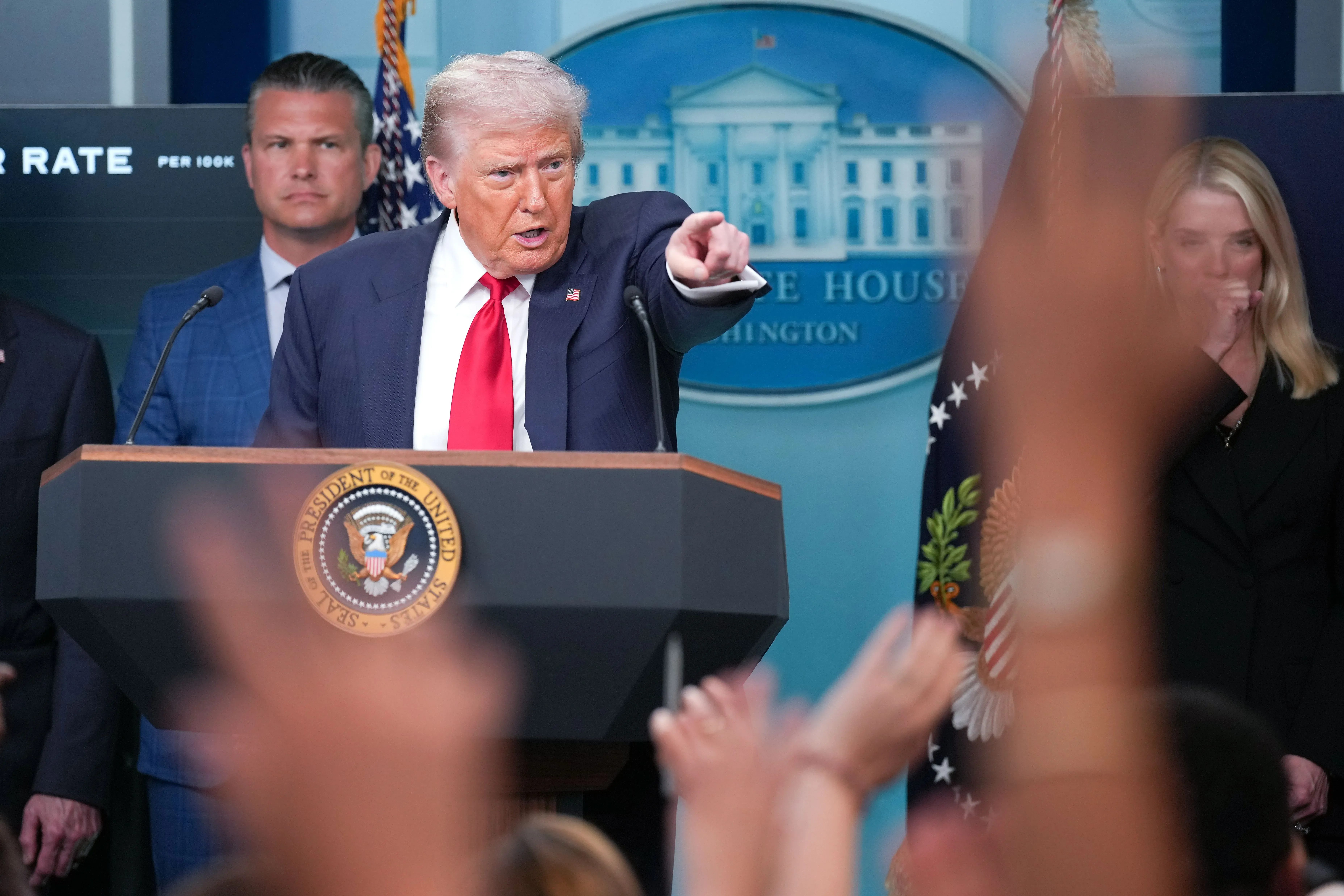 ترامب يتحدث في مؤتمر صحفي بالبيت الأبيض، مشيرًا إلى الحضور، مع ظهور بعض الأيادي المرفوعة في المقدمة.