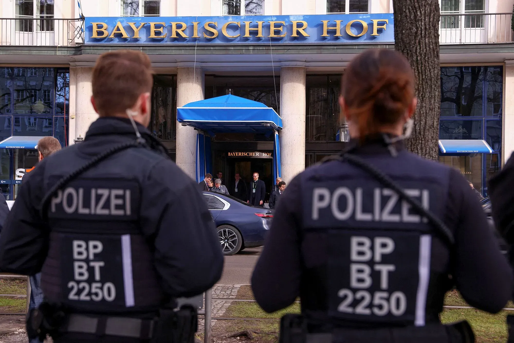 شرطة تحرس مدخل فندق بايريشير هوف في ميونيخ، حيث يُعقد مؤتمر الأمن، مع وجود شخصيات بارزة في الخلفية.