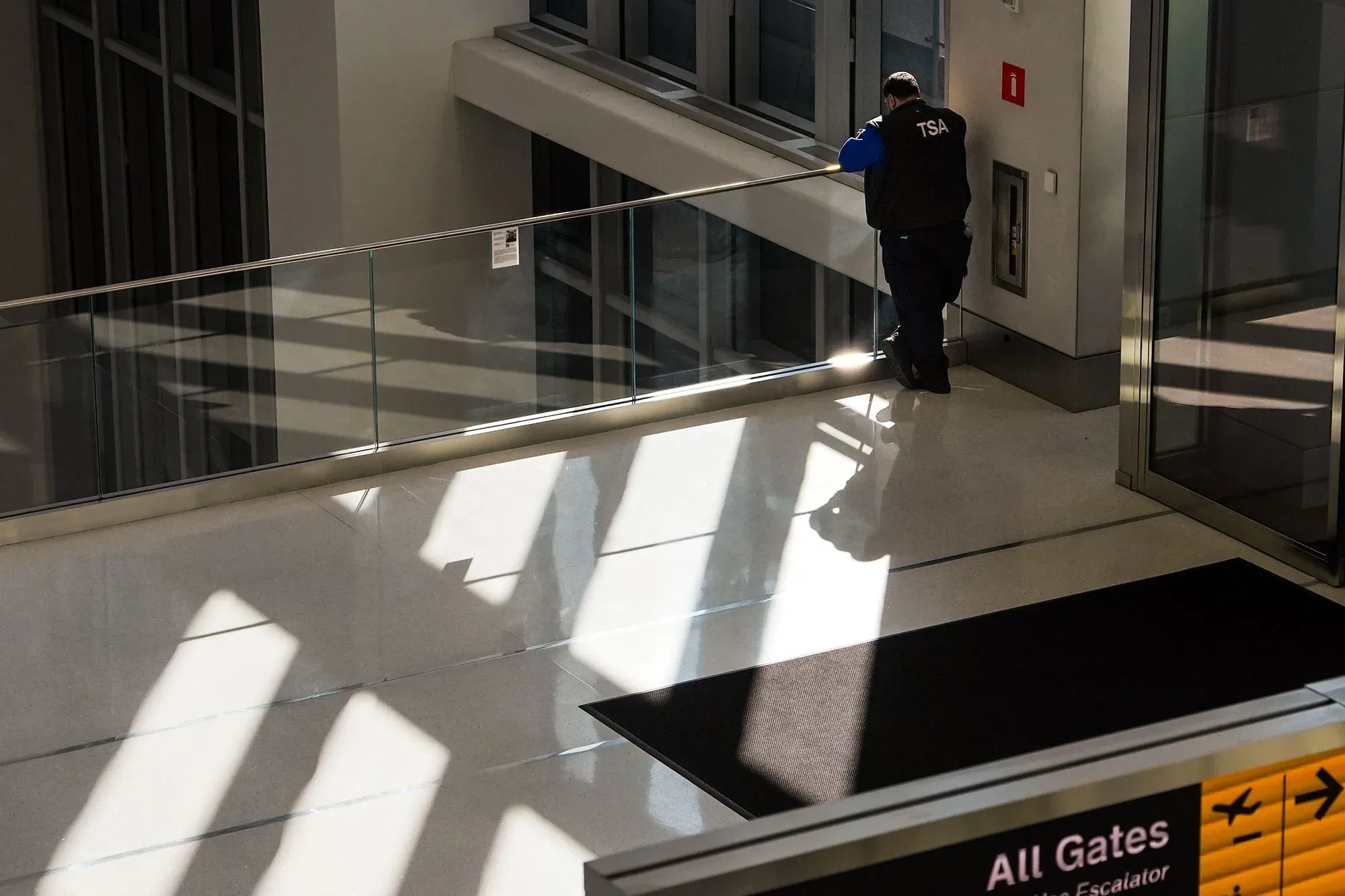 موظف في إدارة أمن النقل يقف في مطار تامبا الدولي، محاطًا بأضواء وظلال، مع شعور بالقلق بسبب تأخر الرواتب.