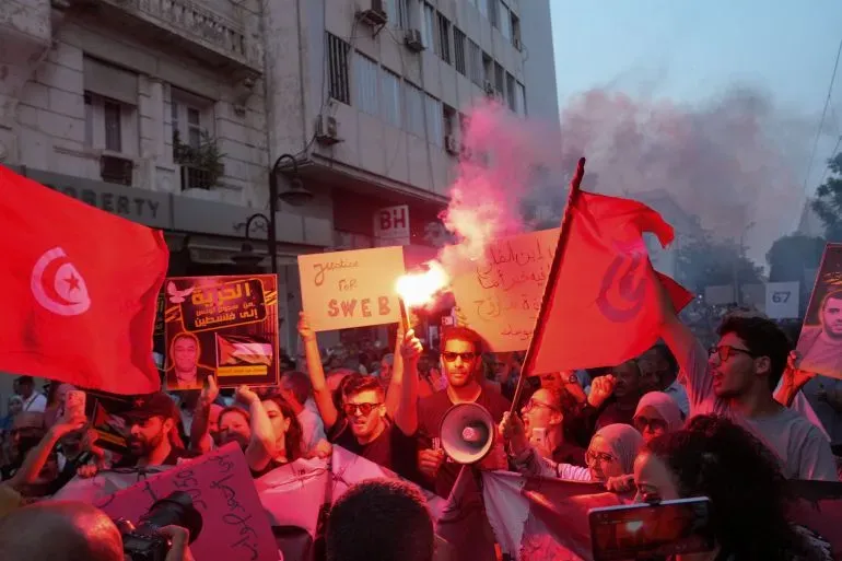 محتجون تونسيون يحملون لافتات وأعلامًا، مع إشعال الألعاب النارية، في مظاهرة ضد حكم قيس سعيد، مطالبين بالديمقراطية والإفراج عن المعتقلين السياسيين.