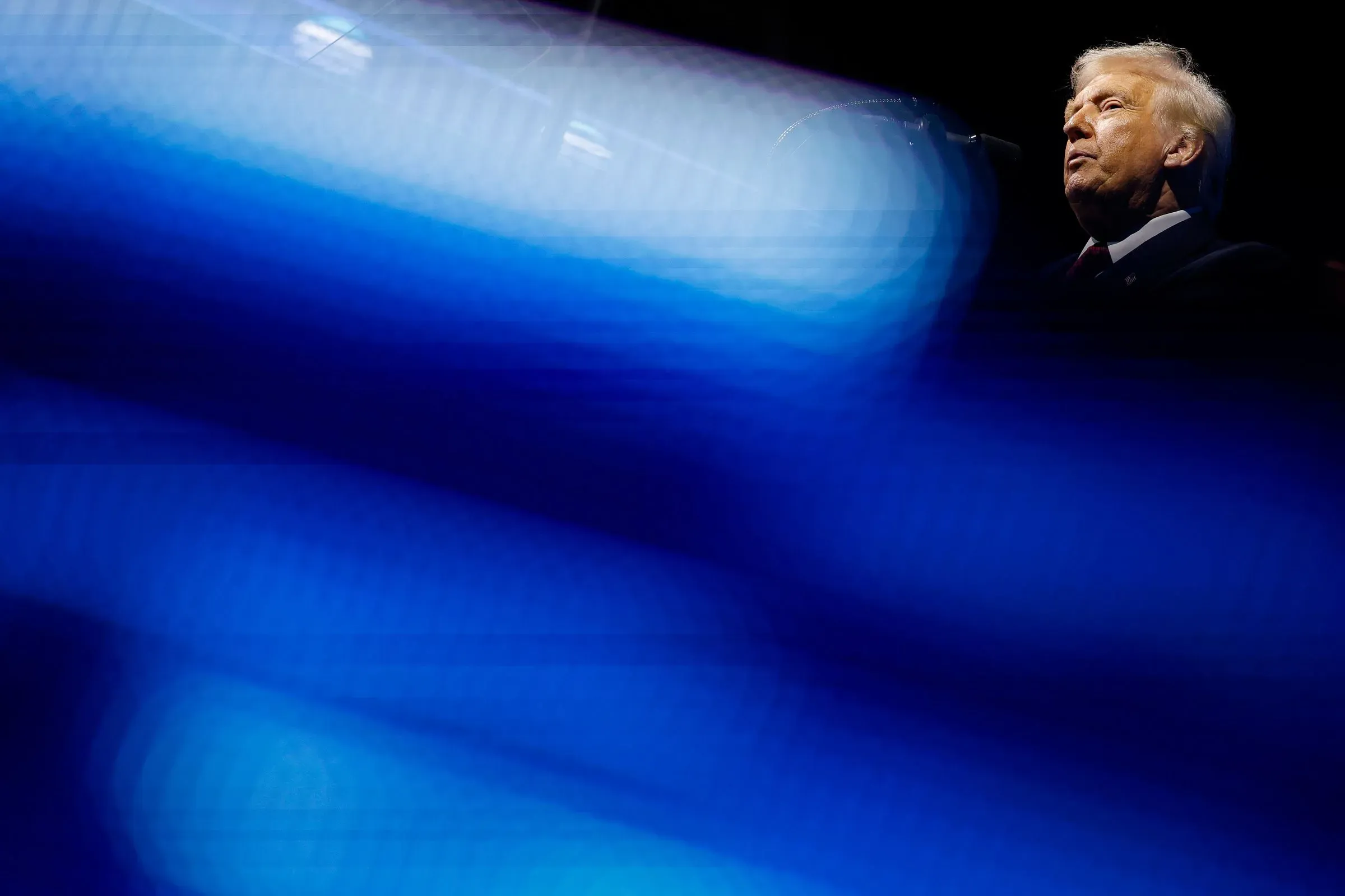 صورة لدونالد ترامب في تجمع سياسي، يظهر فيها بوضوح خلف ميكروفون، مع تأثيرات ضوئية زرقاء تعكس الأجواء الحماسية.