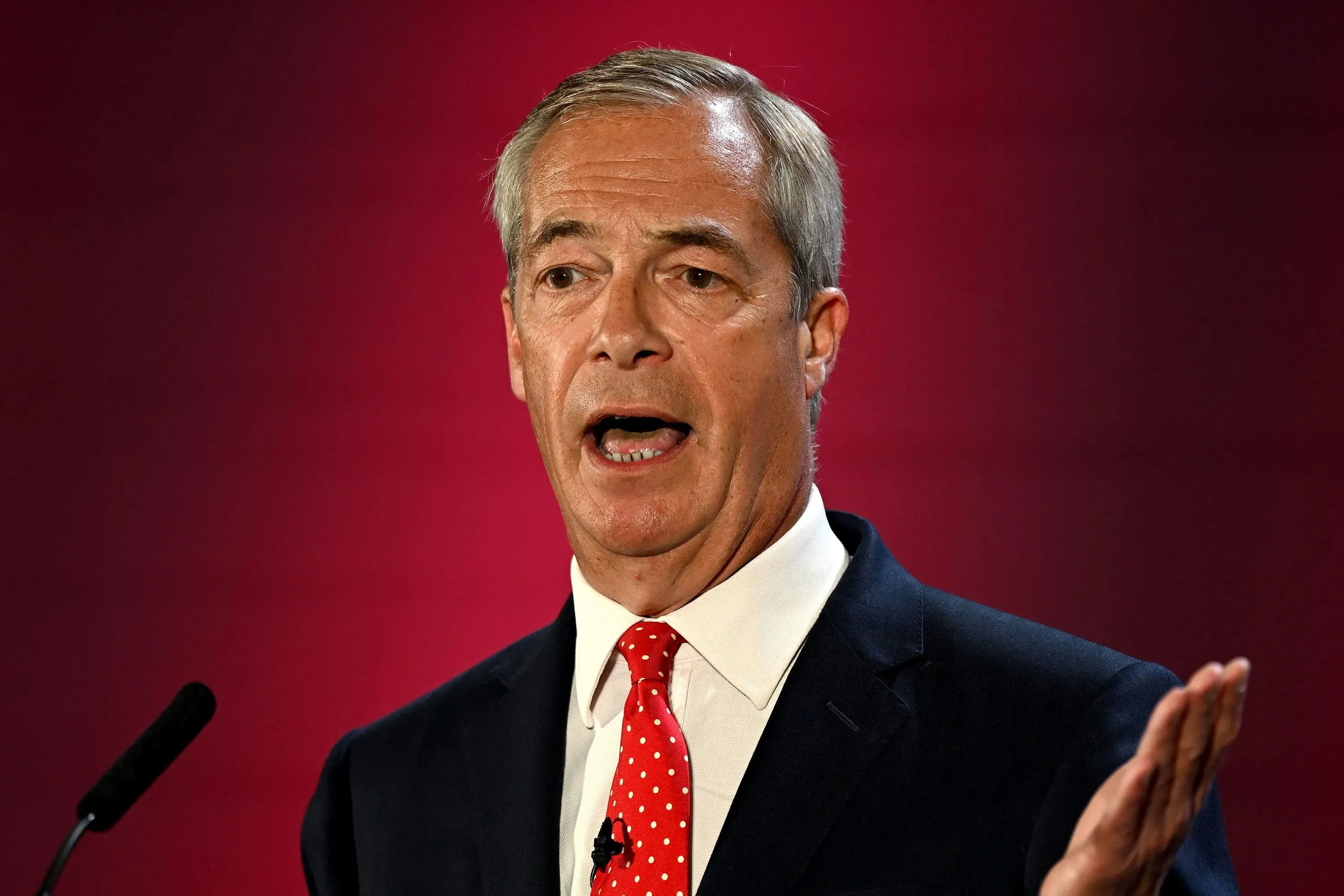 نايجل فاراج يتحدث في مؤتمر صحفي حول خطط ترحيل طالبي اللجوء في المملكة المتحدة، مع خلفية حمراء تعكس التوتر السياسي.