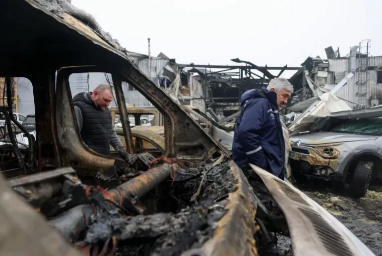 رجال يتفقدون الأضرار في موقع تعرض لهجوم، حيث تظهر سيارات مدمرة ومبانٍ متضررة في الخلفية، مما يعكس آثار النزاع المستمر في المنطقة.
