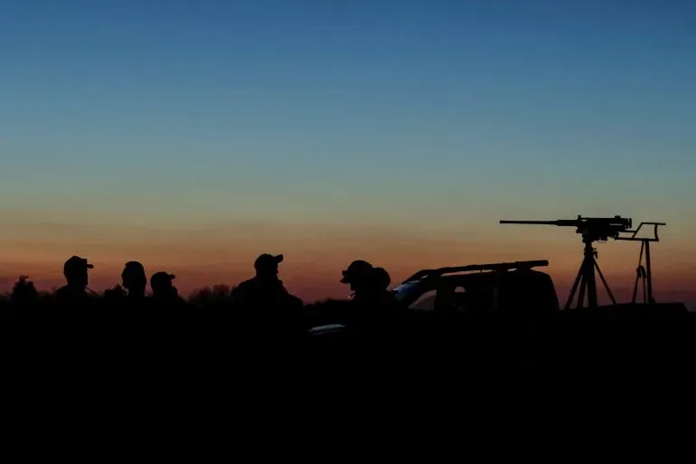 مجموعة من الأشخاص يقفون silhouetted أمام غروب الشمس، مع وجود سلاح مضاد للطائرات مثبت على قاعدة، مما يعكس تصاعد التوترات العسكرية في الصراع الأوكراني.